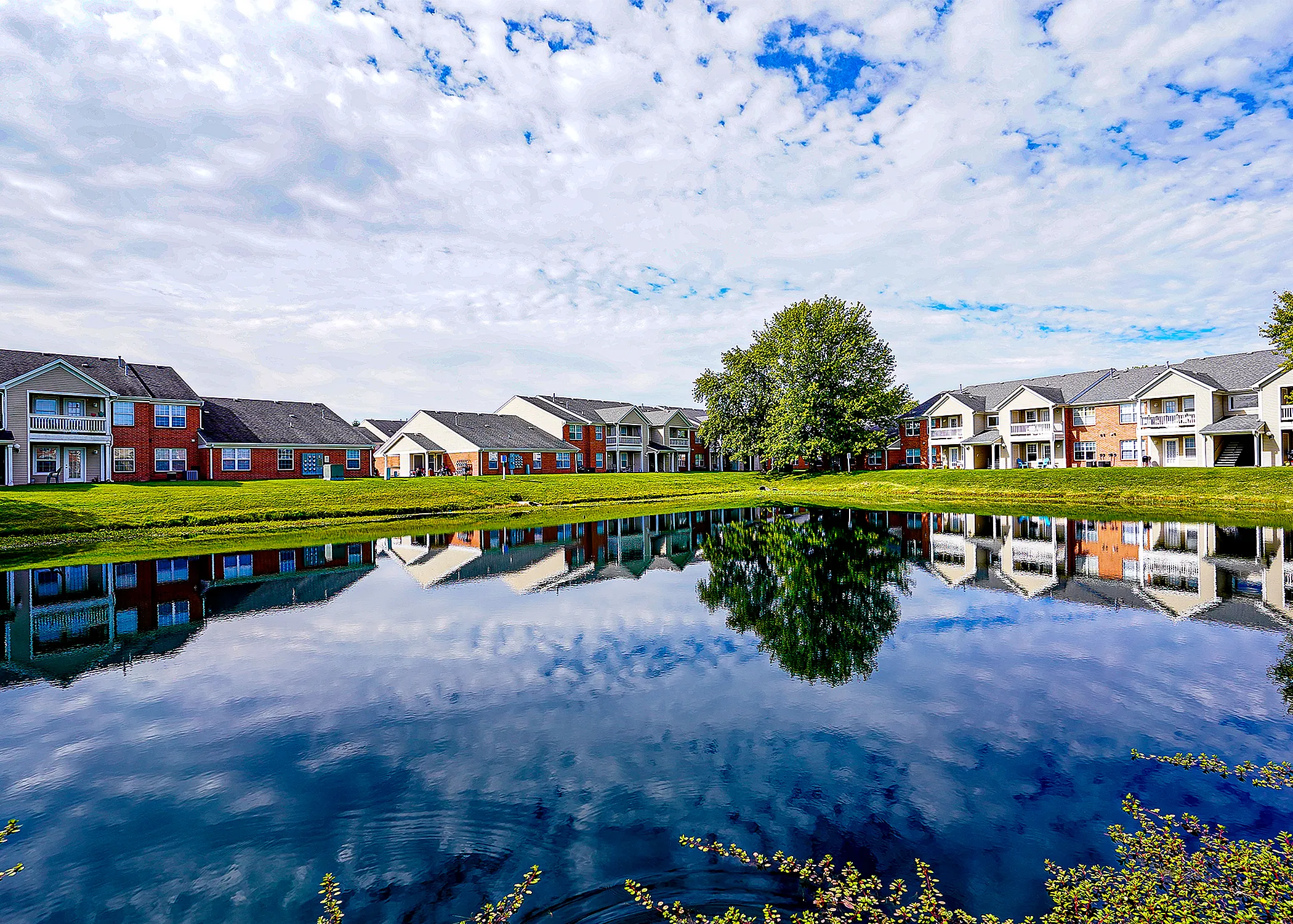 Charming apartment complex with scenic pond reflection under a vibrant sky, surrounded by lush greenery and modern architecture.