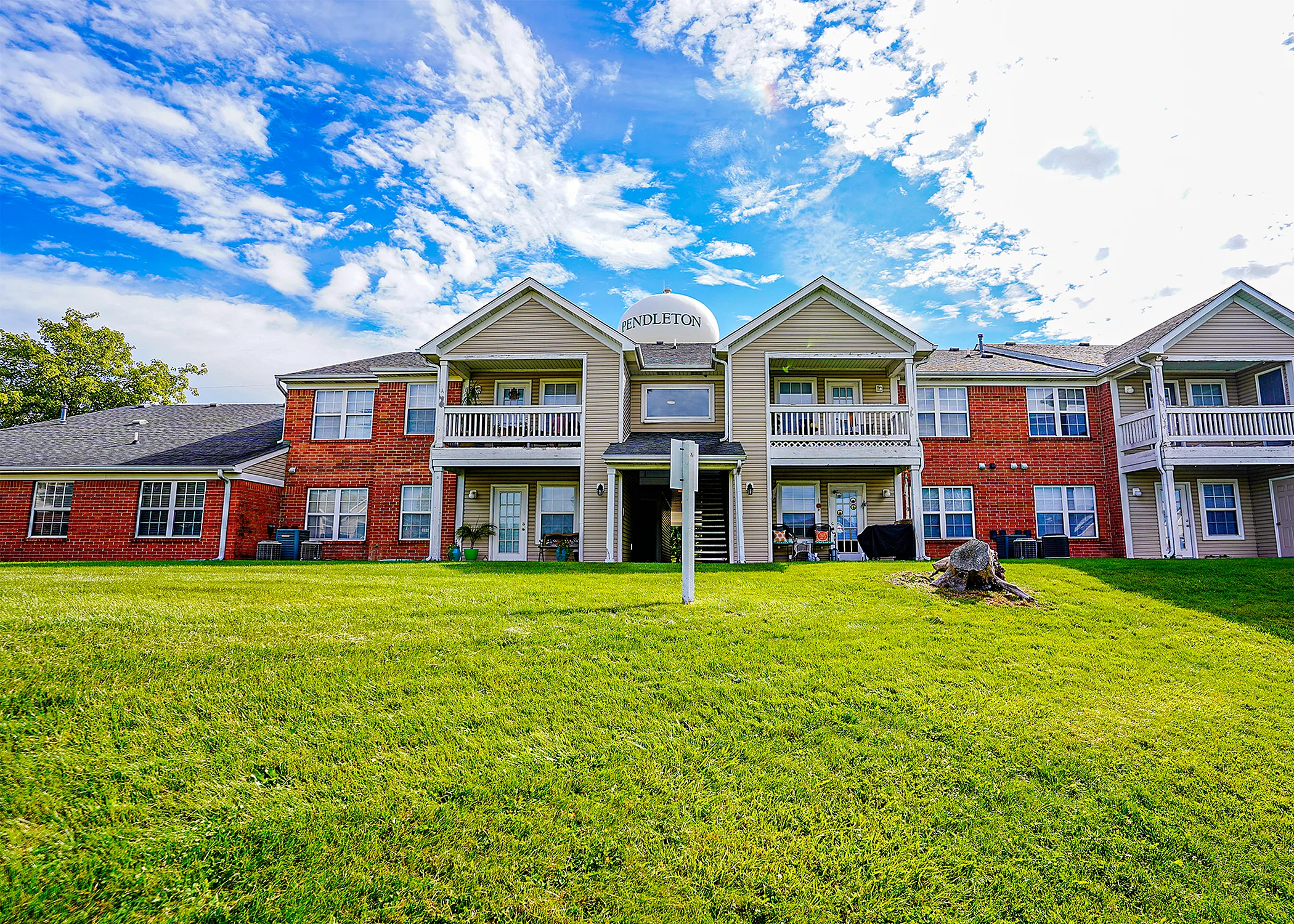 Charming apartment complex with red brick and beige siding, featuring balconies and lush green lawns under a vibrant blue sky.