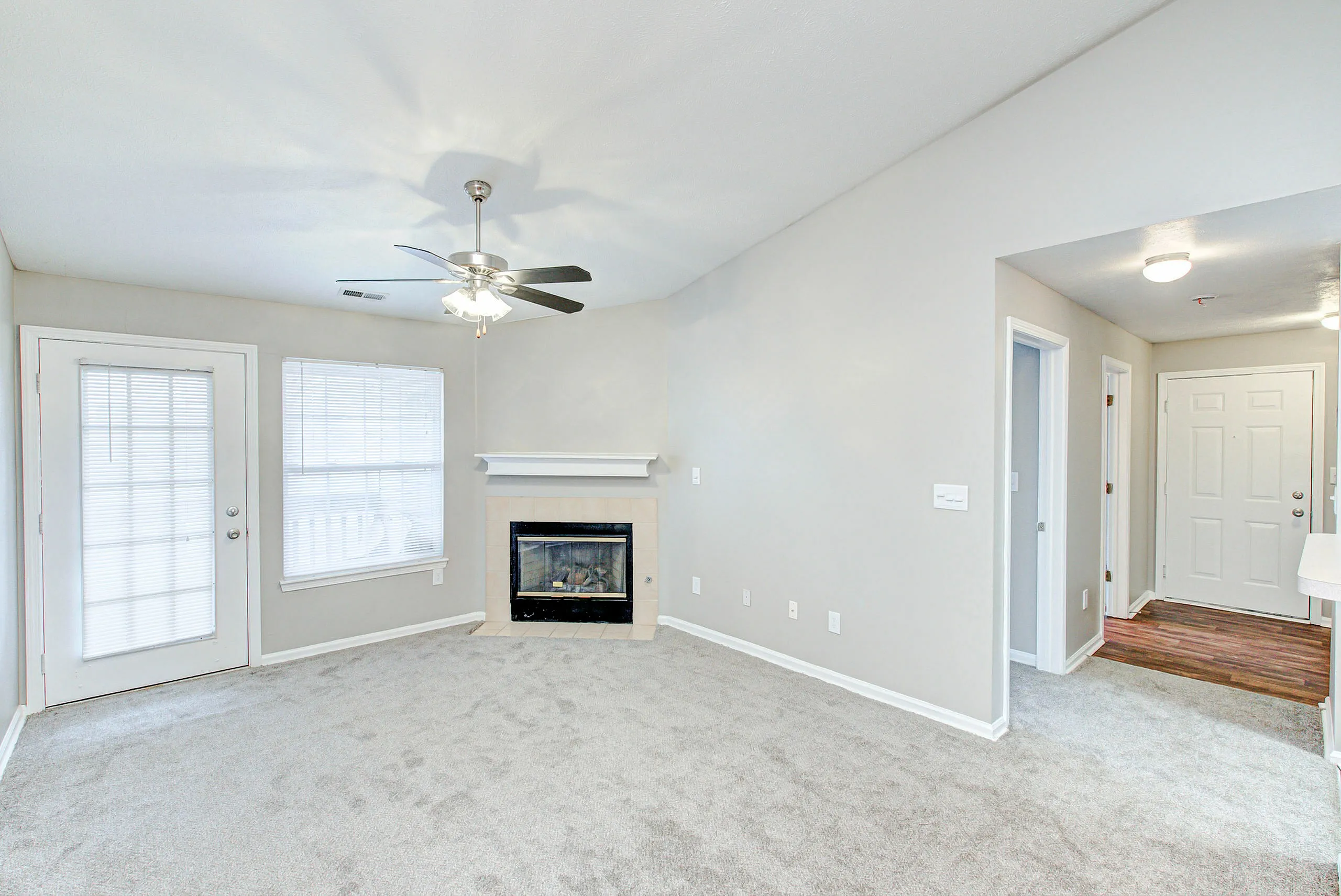 Bright living room featuring plush carpet, modern ceiling fan, and a cozy fireplace, with natural light streaming through glass doors and windows.