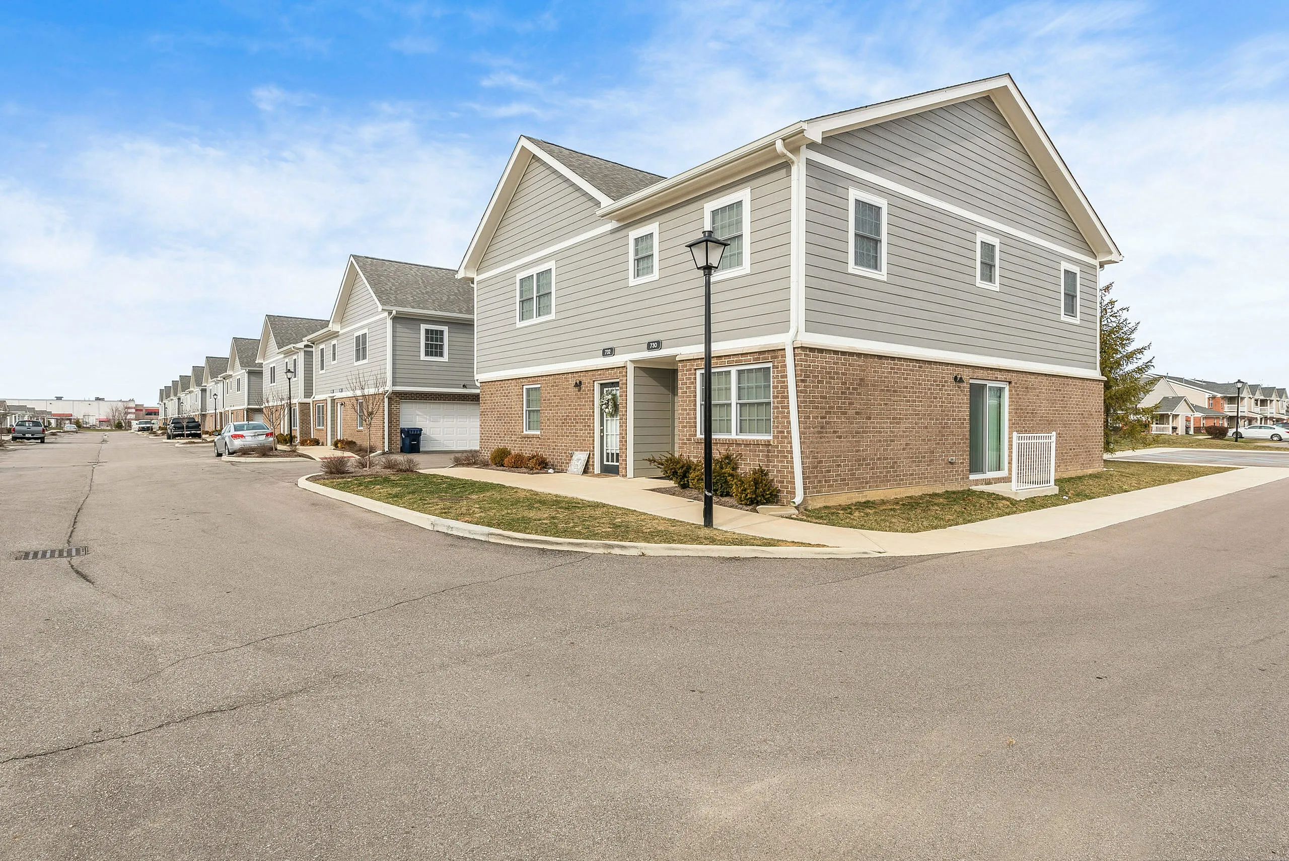 Charming two-story townhouse with modern gray siding and brick facade, nestled in a peaceful suburban neighborhood under a bright blue sky.