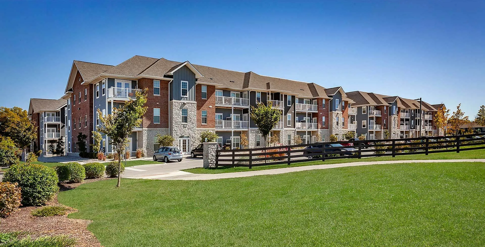 Elegant apartment complex with modern brick facade and spacious balconies, set in a lush green landscape under a clear blue sky.