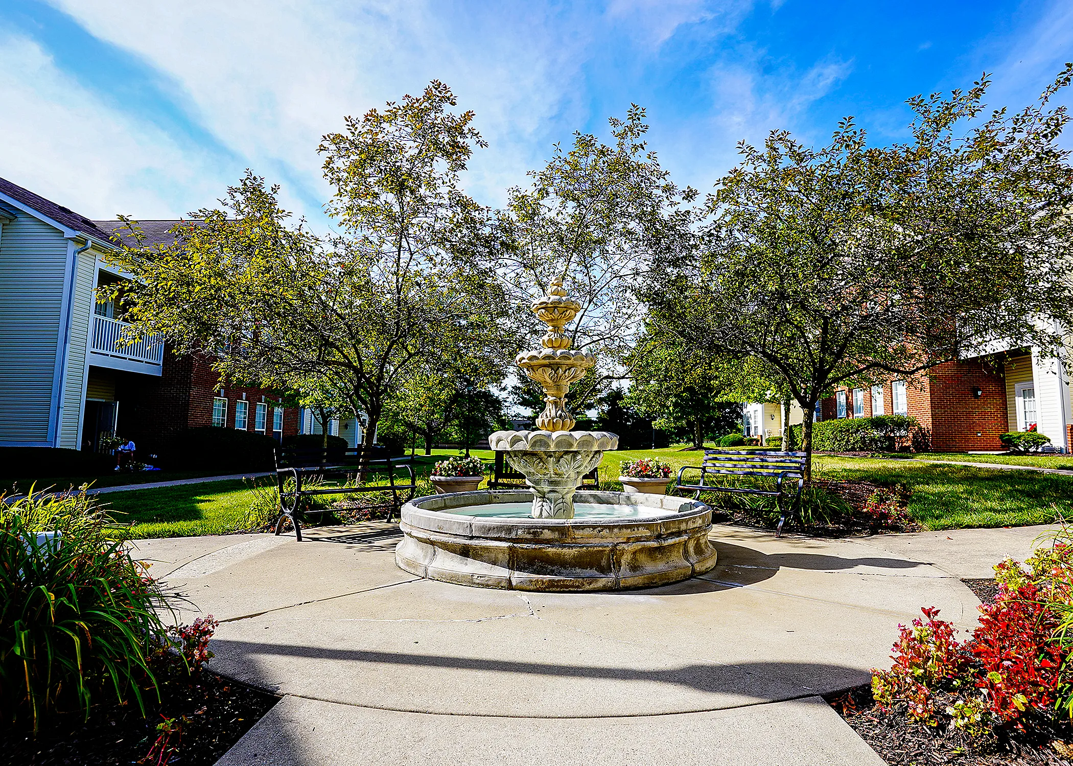 Charming courtyard view with a beautiful stone fountain surrounded by lush greenery and benches, perfect for relaxation in a serene apartment complex.