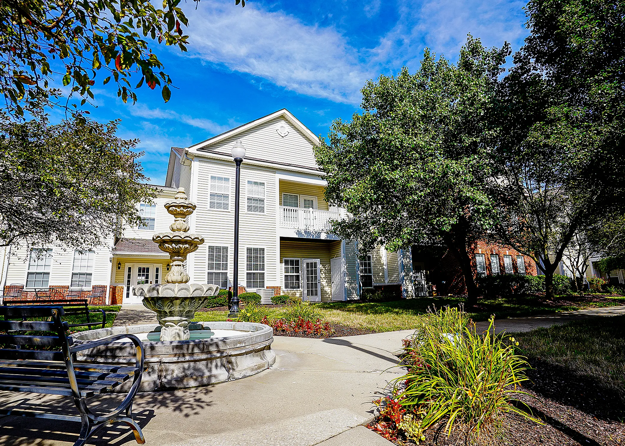 Charming apartment building surrounded by lush greenery, featuring a classic stone fountain and inviting pathways under a clear blue sky.