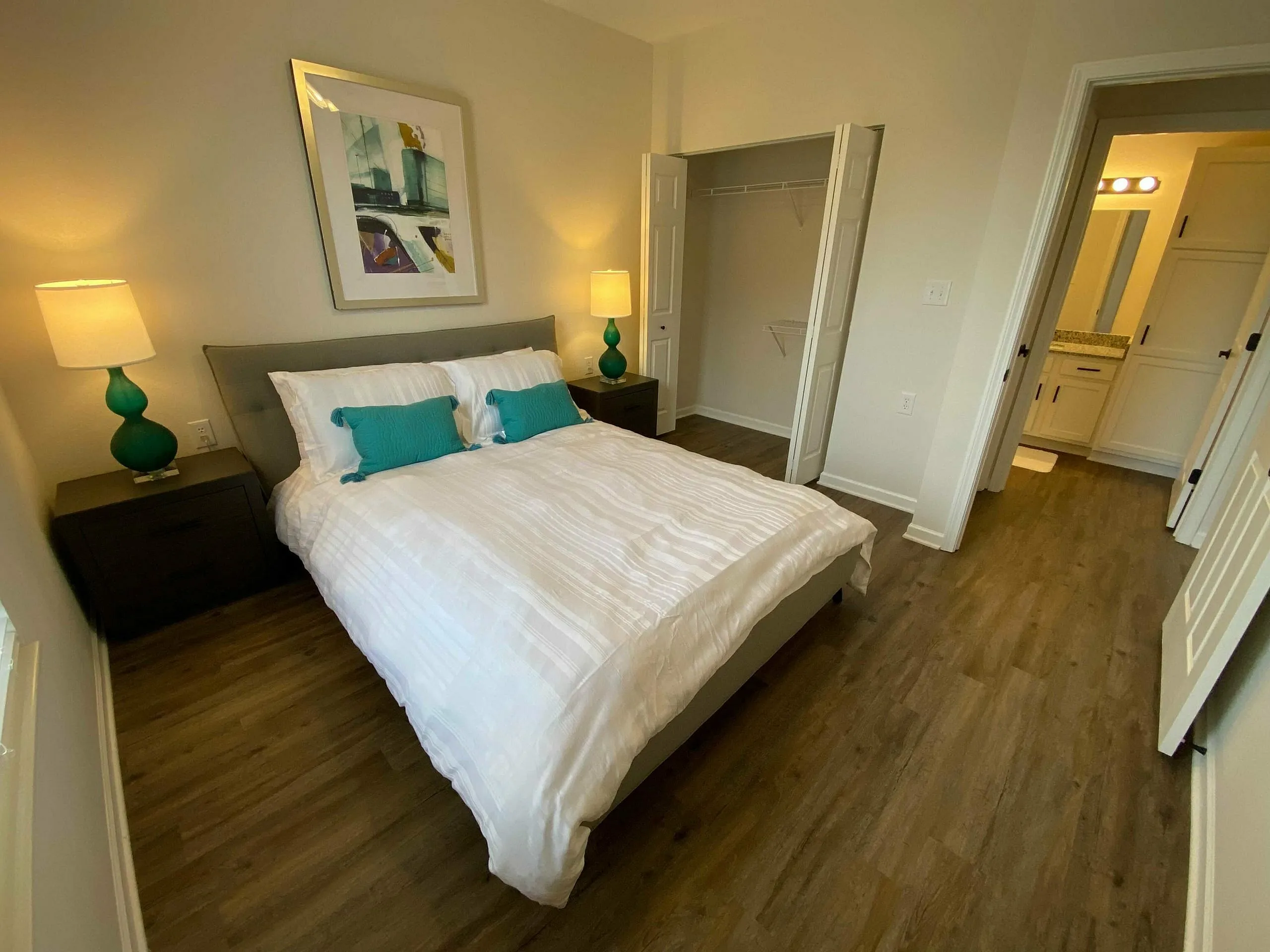 Inviting bedroom with modern decor, featuring a cozy bed with teal accents, elegant artwork, and warm lighting, leading to an en-suite bathroom.
