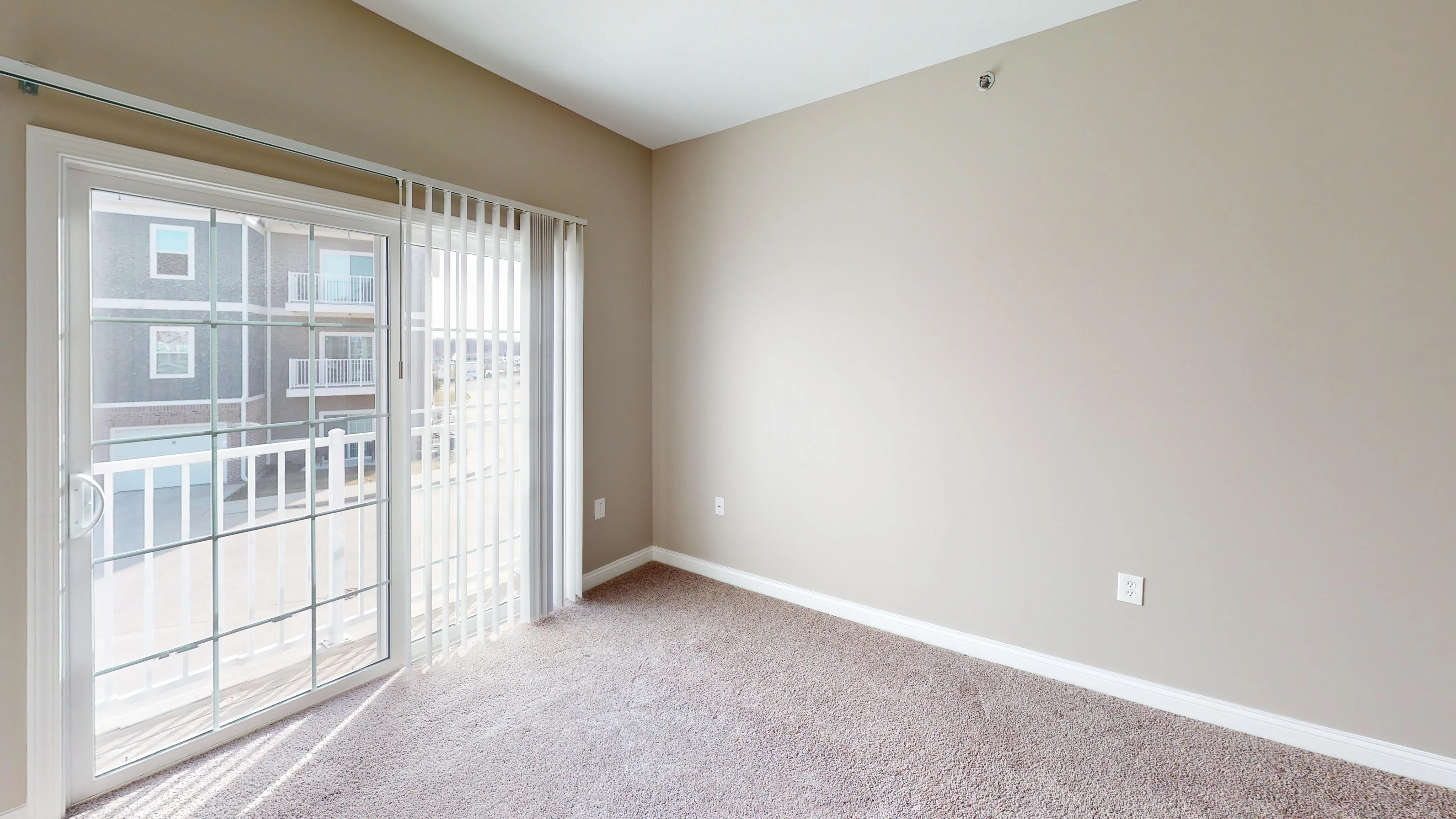 Bright and inviting room with plush carpet, large window, and modern white blinds, offering a view of neighboring balconies.