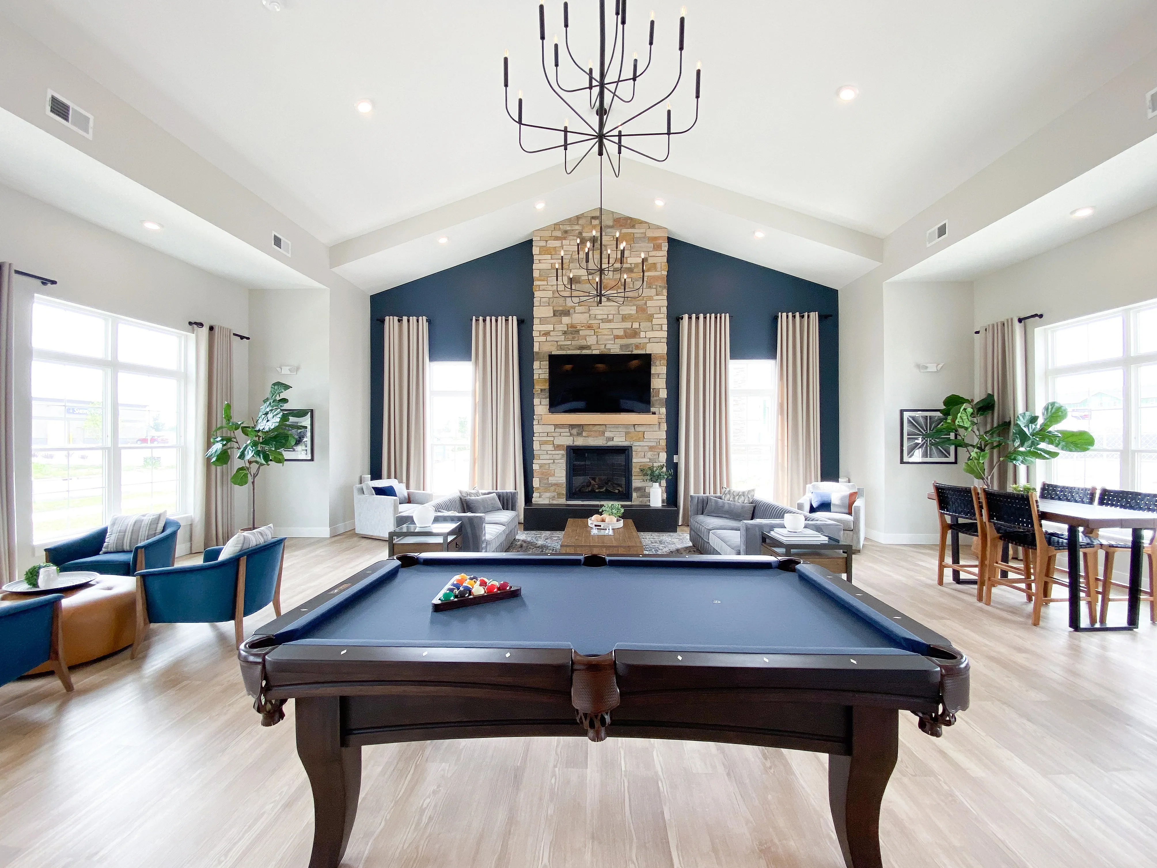Spacious and elegant living room featuring a luxurious pool table, cozy seating area with a stone fireplace, high ceilings with a chandelier, and large windows allowing natural light to flood the space.