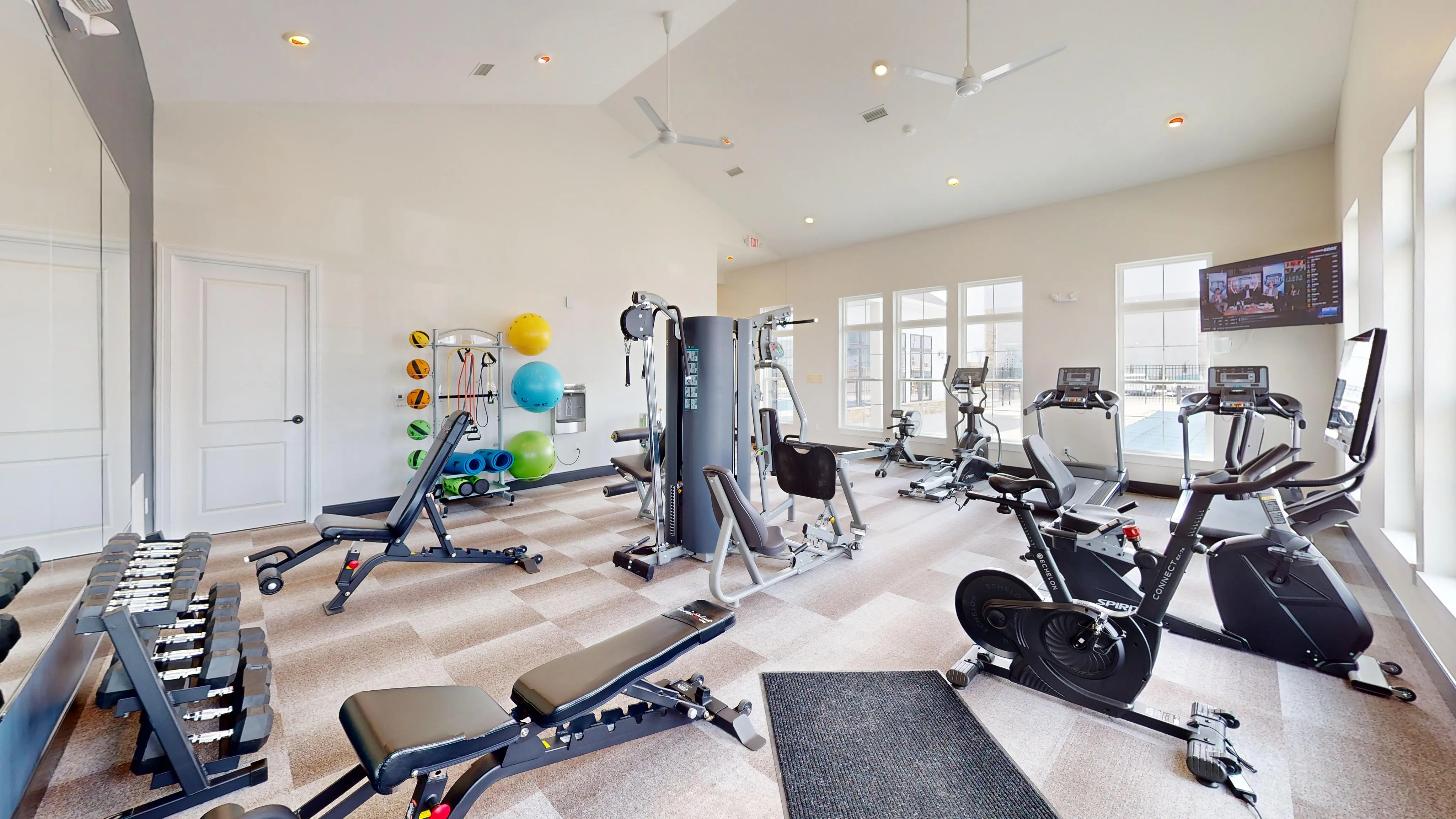 State-of-the-art fitness center with modern equipment, including treadmills, exercise bikes, and weight machines against a backdrop of bright, sunlit windows.