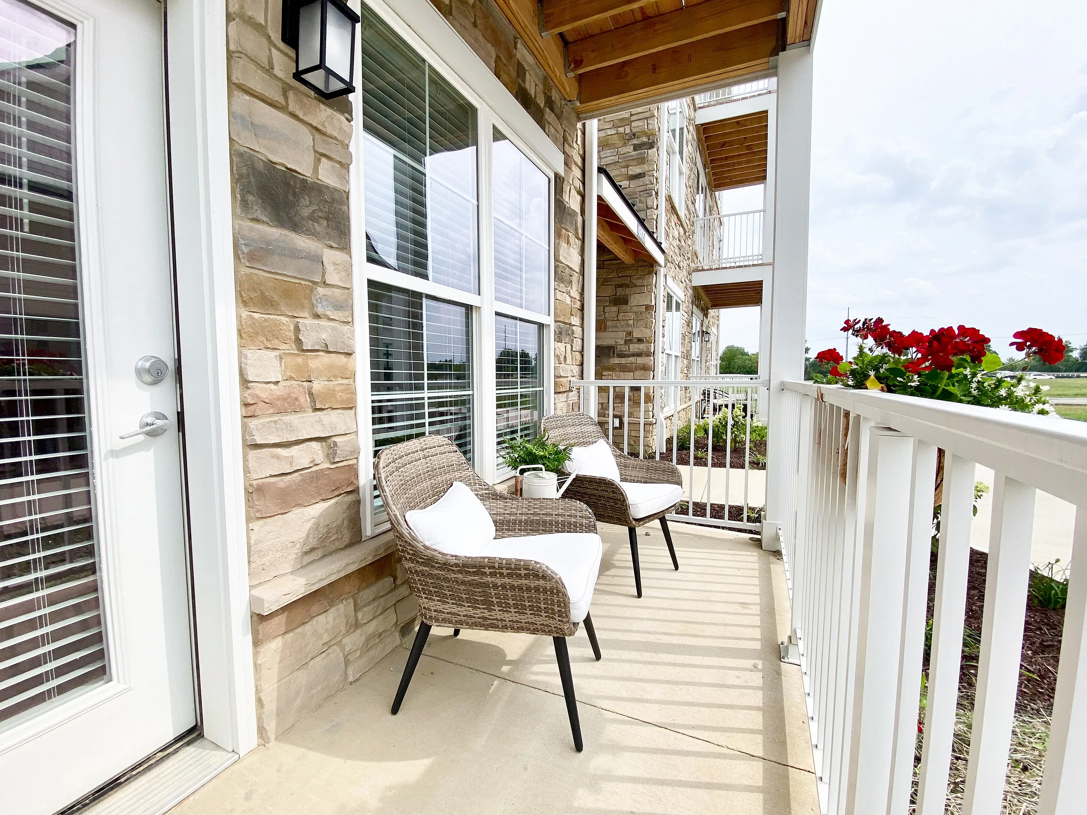 Charming apartment patio with cozy wicker chairs, vibrant red flowers, and elegant stone siding, offering a serene outdoor retreat.