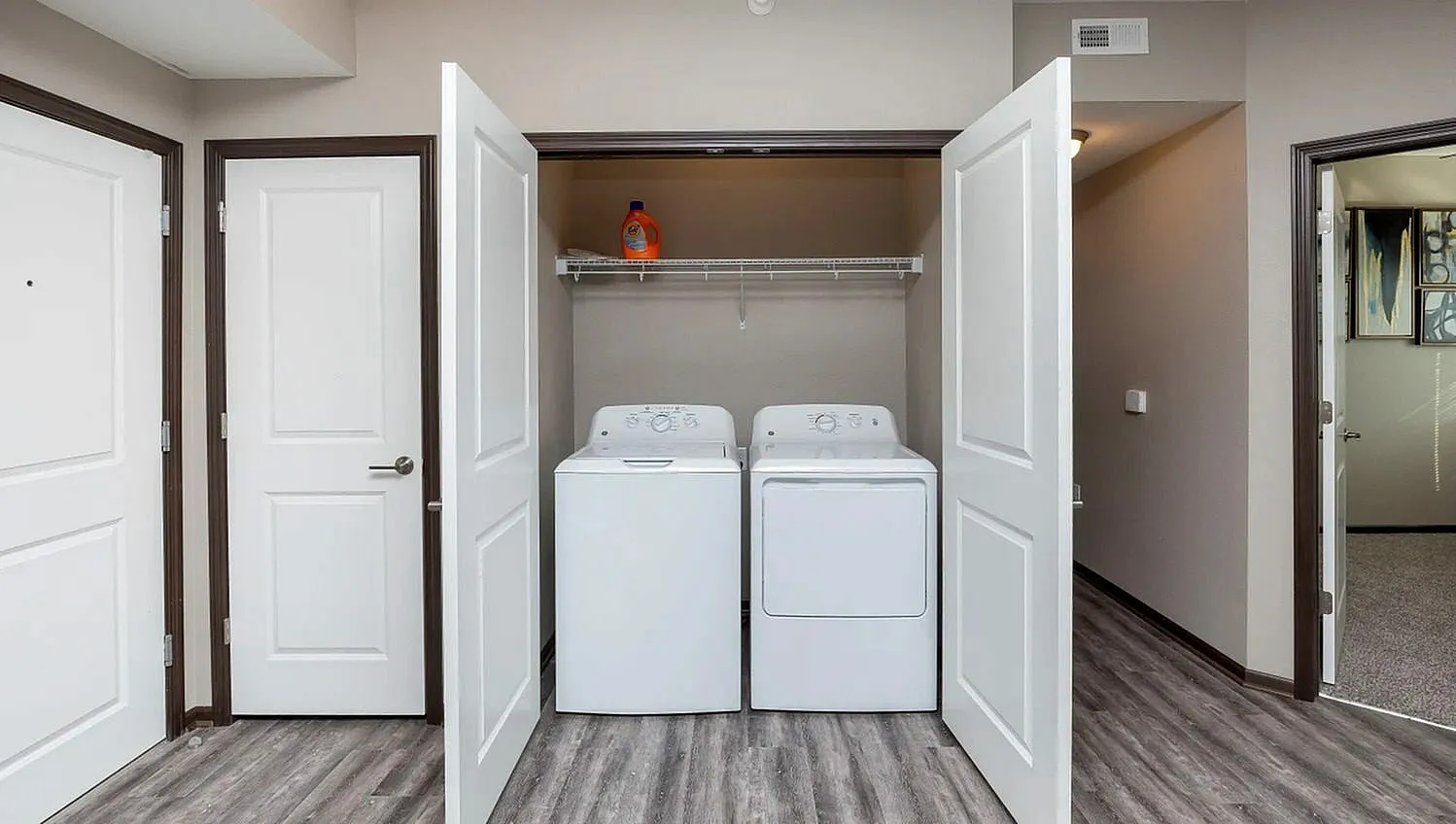 Modern apartment laundry nook with white washer and dryer, sleek laminate flooring, and double doors for convenience.