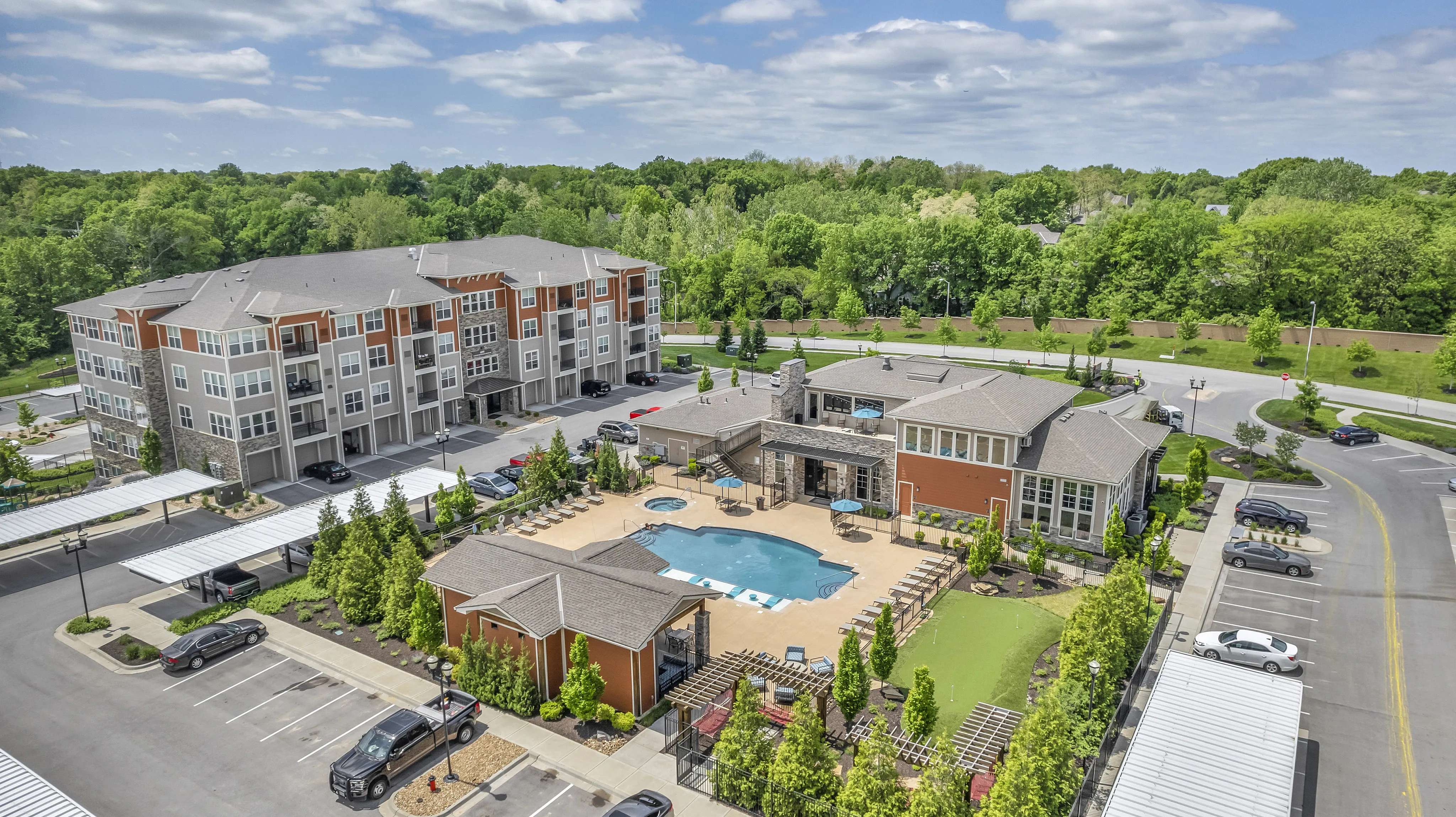 Luxurious apartment complex with modern architecture, featuring a central swimming pool, lush greenery, and ample parking against a scenic wooded backdrop.