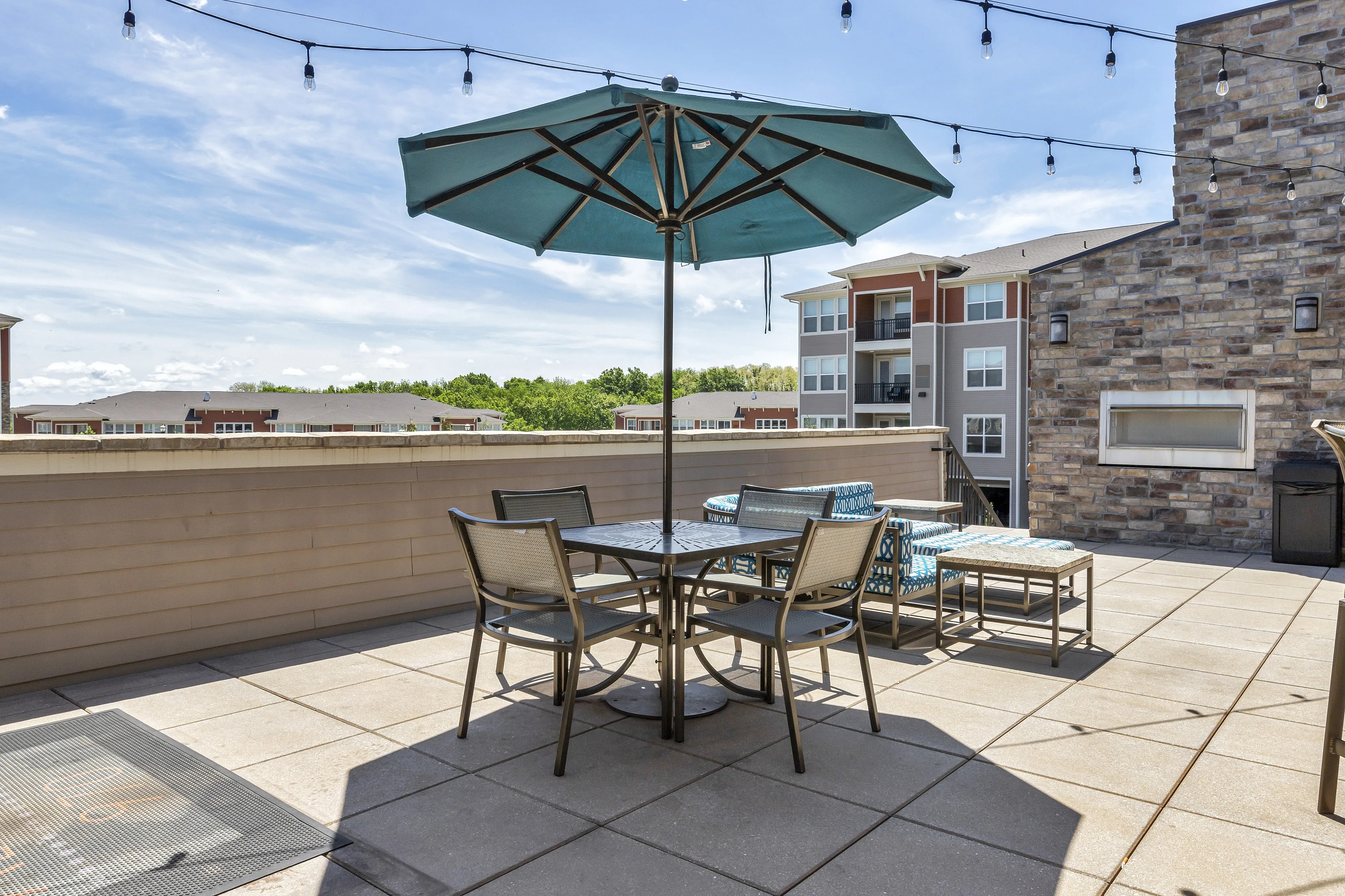 Charming rooftop patio with stylish outdoor seating and umbrella, surrounded by modern apartment buildings under a clear blue sky.