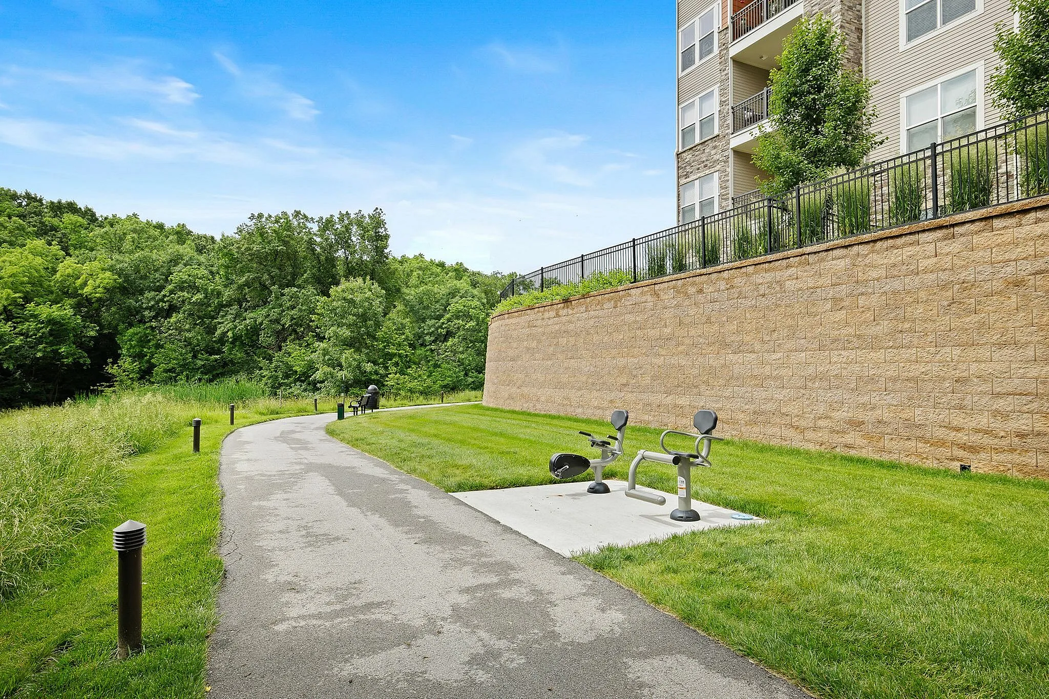 Inviting outdoor area with a serene walking path and modern fitness equipment, nestled alongside lush green trees and a well-maintained apartment complex.
