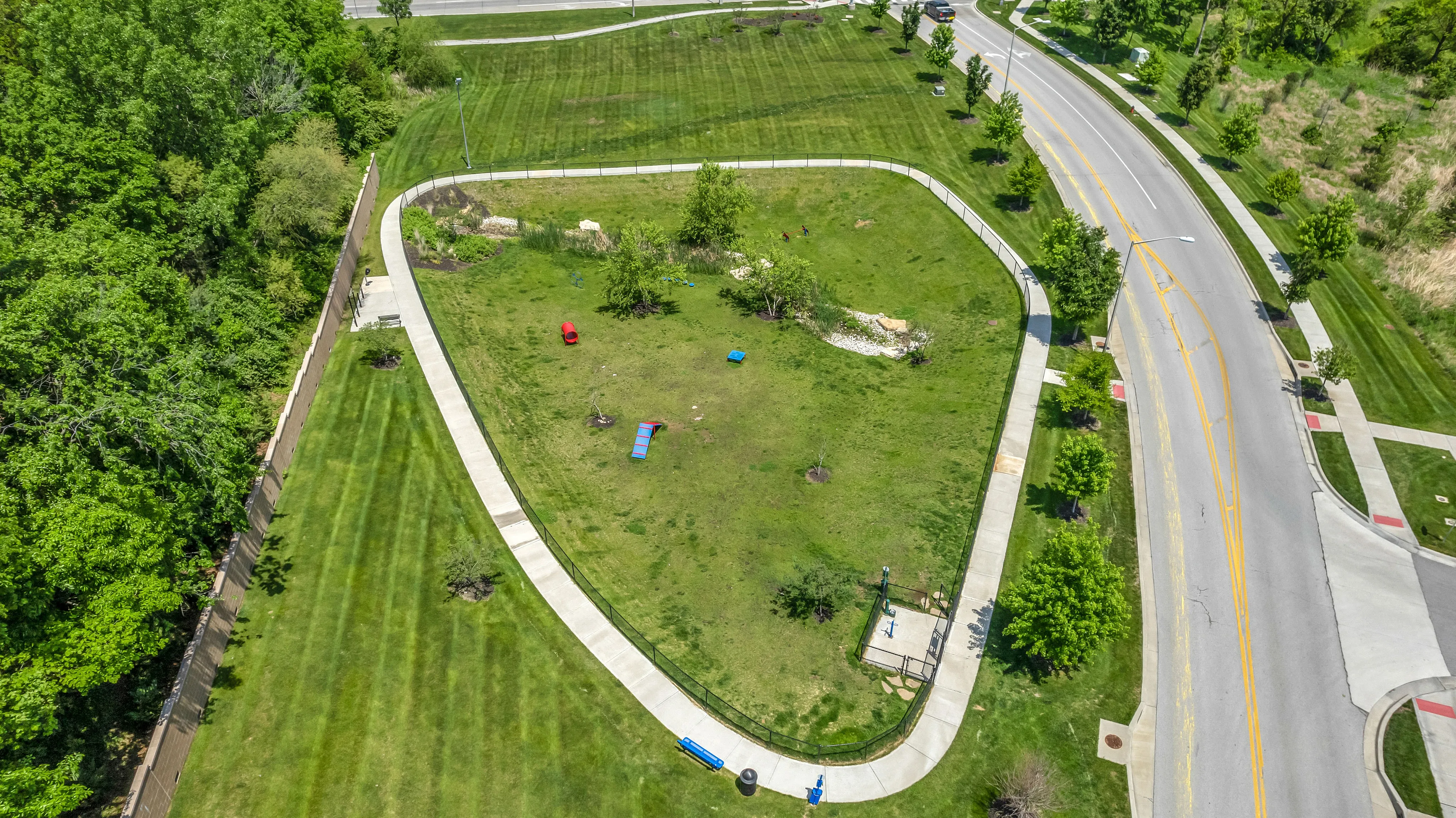 Expansive aerial view of a lush, triangle-shaped dog park with vibrant green grass, walking paths, and surrounding trees.