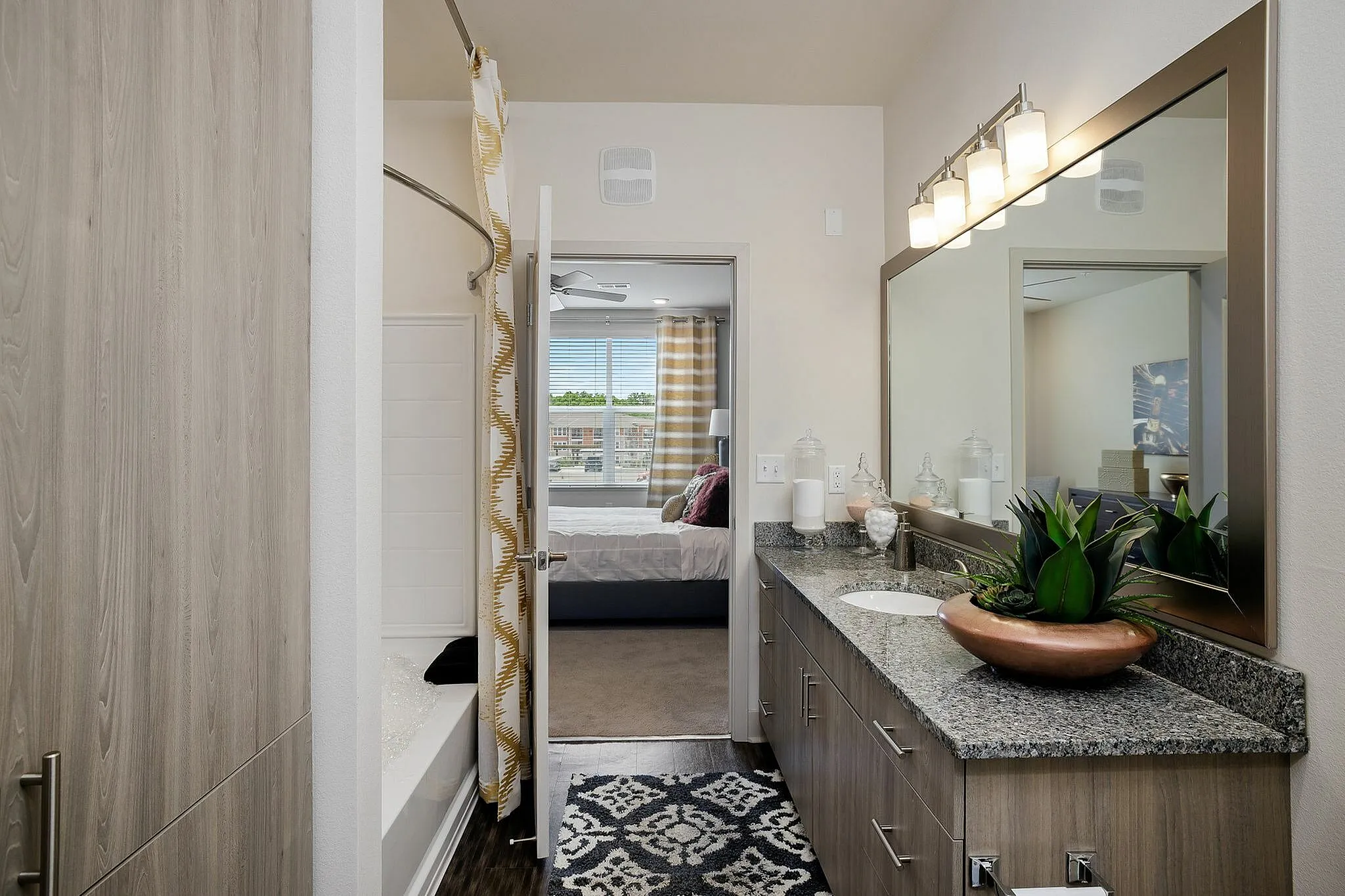 Elegant bathroom view featuring a sleek granite countertop, large mirror with modern lighting, and stylish plant decor leading to a cozy bedroom.