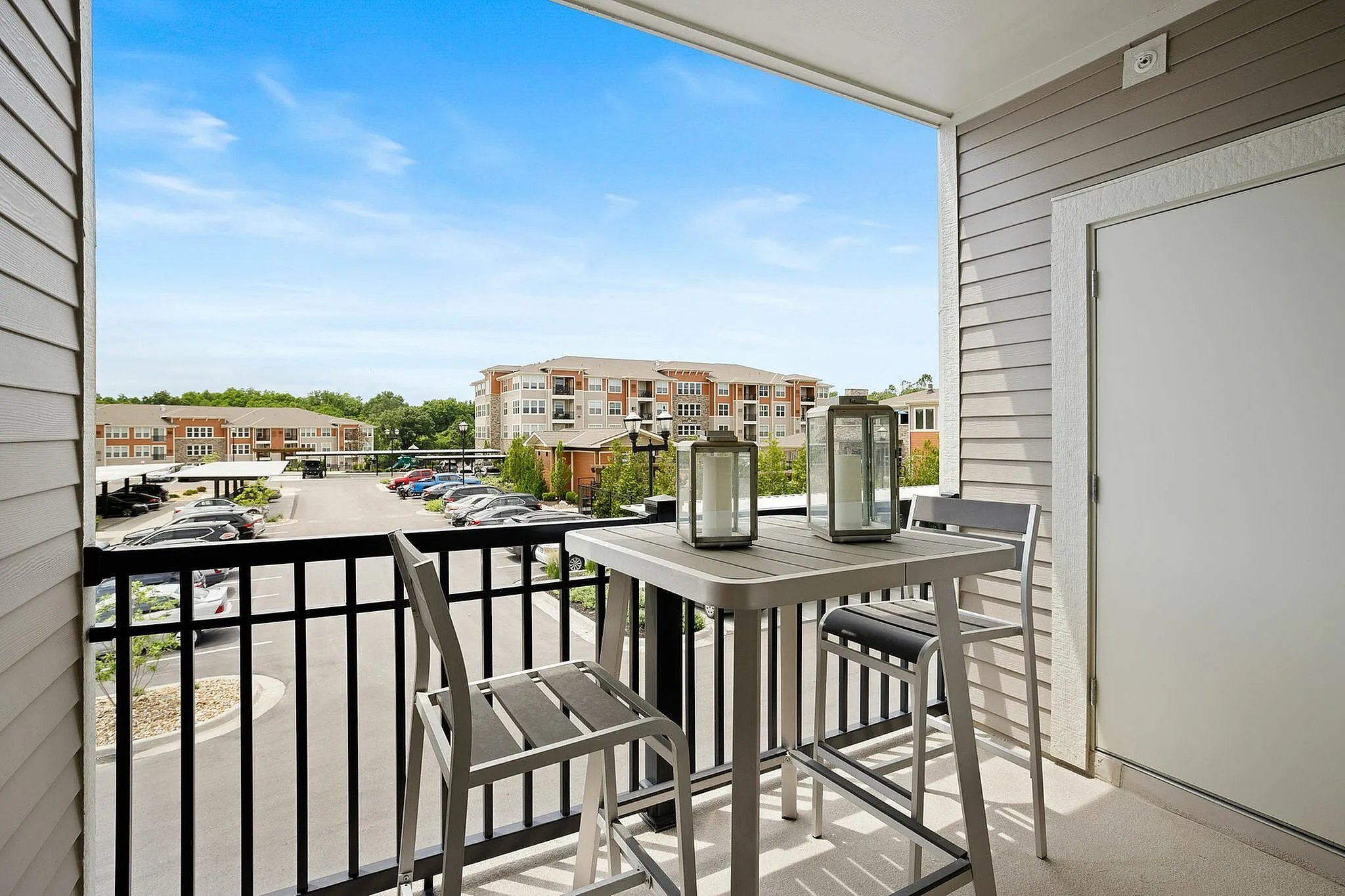 Charming balcony with cozy seating, offering scenic views of modern apartment complex and parking area under a bright blue sky.