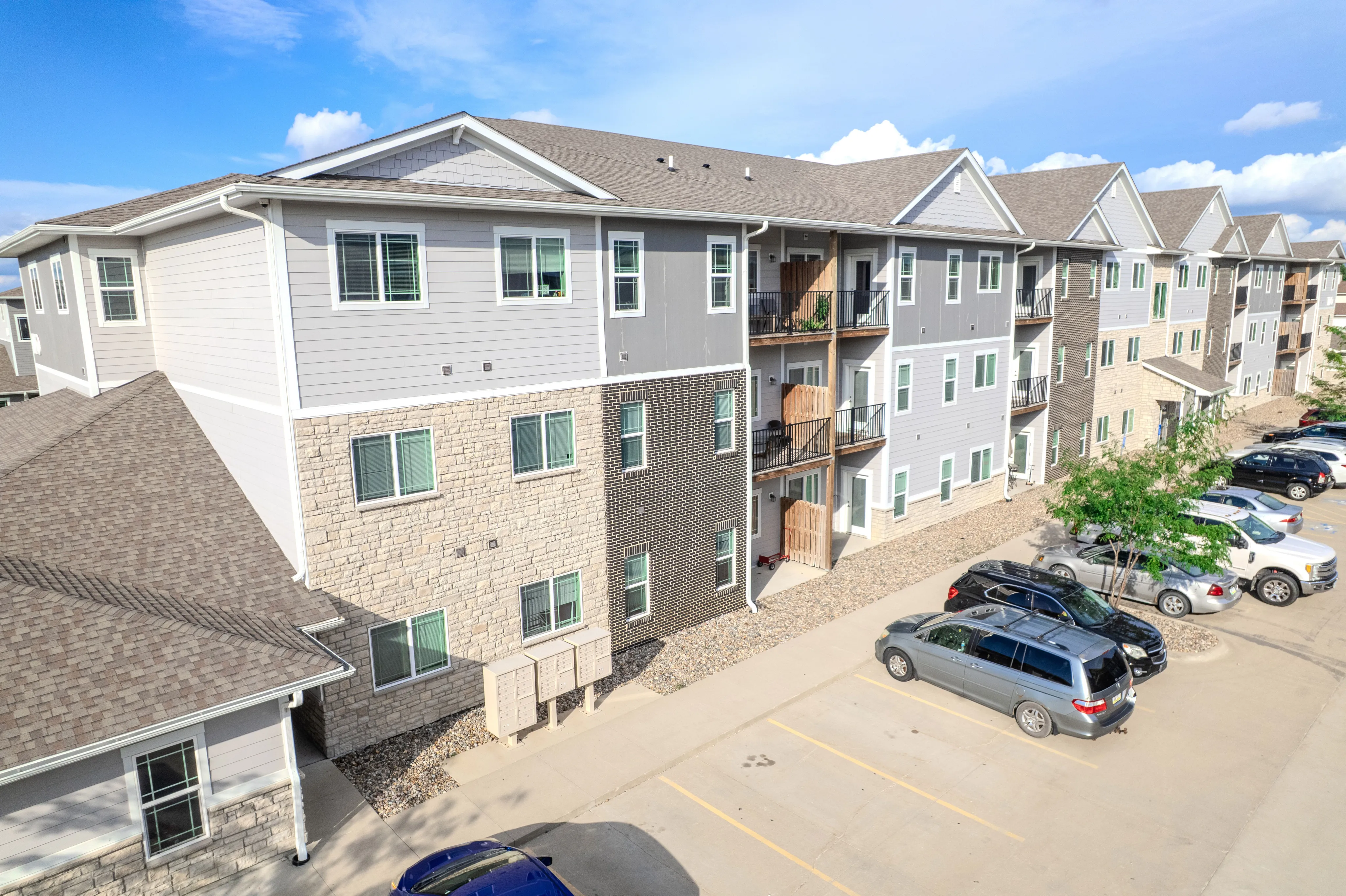 Modern apartment complex with three stories, featuring sleek exterior design, spacious balconies, and ample parking under a bright blue sky.
