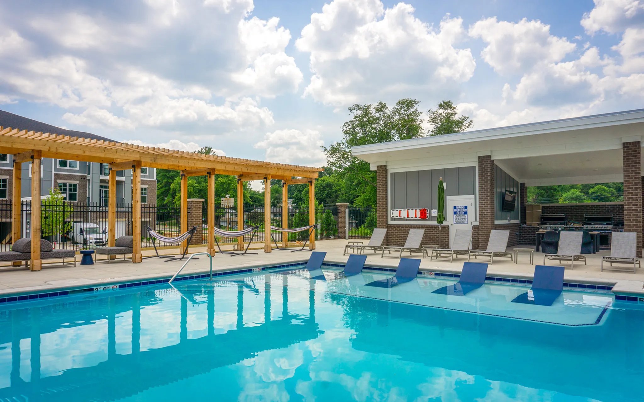 Luxurious apartment pool area featuring a sparkling blue pool, comfy loungers, stylish hammocks, and a chic outdoor kitchen under a sunny sky.