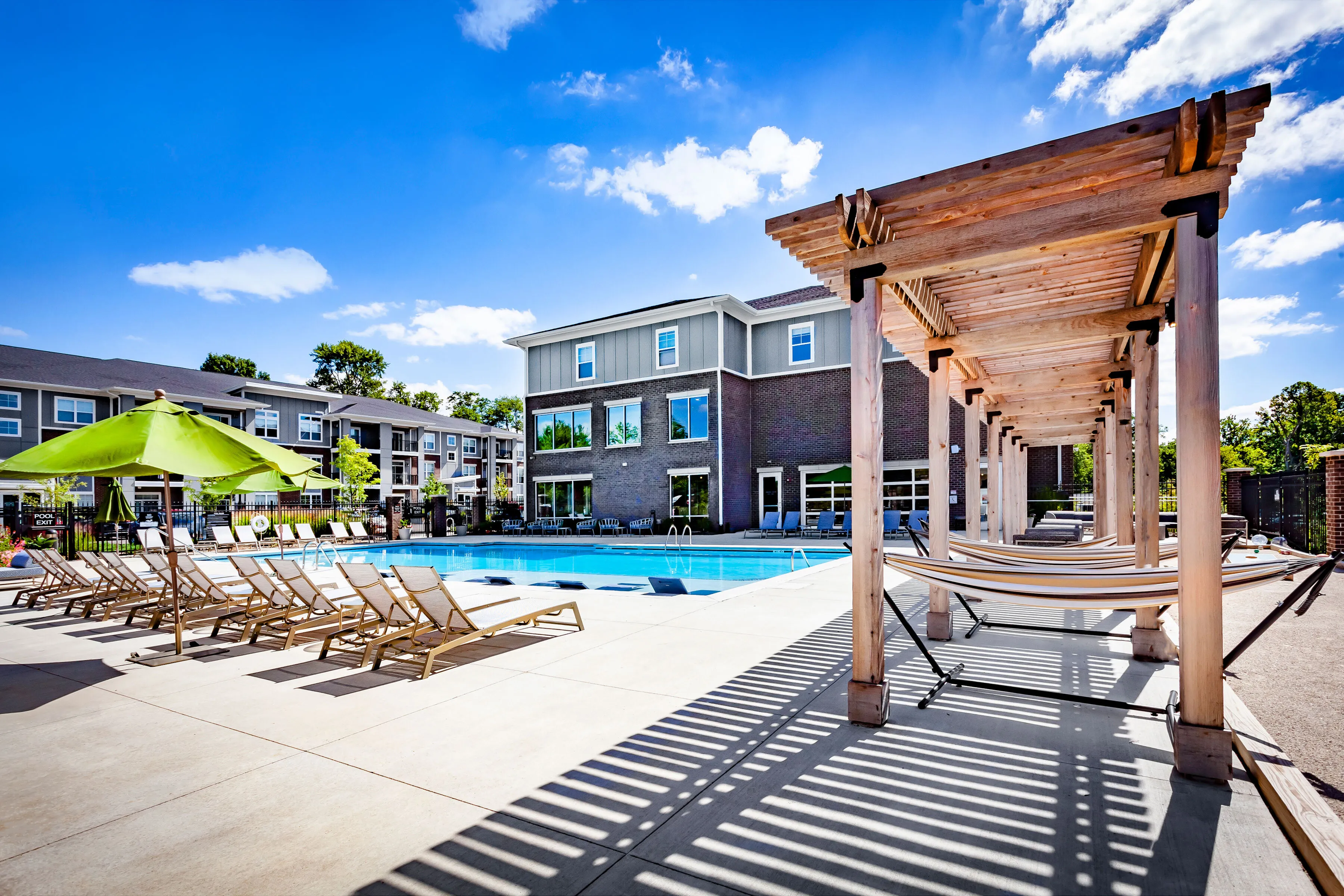Modern apartment complex with a luxurious outdoor pool area, featuring comfortable sun loungers, green umbrellas, and stylish pergolas under a clear blue sky.