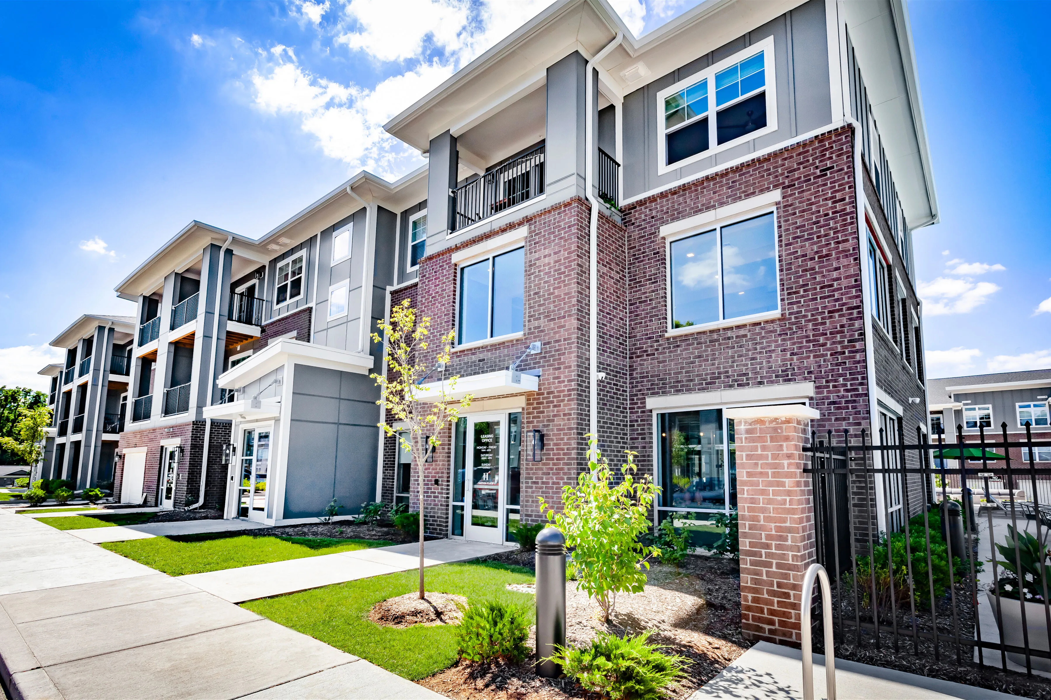 Modern apartment complex with stylish brick facade, large windows, and well-maintained landscaping under a bright blue sky.