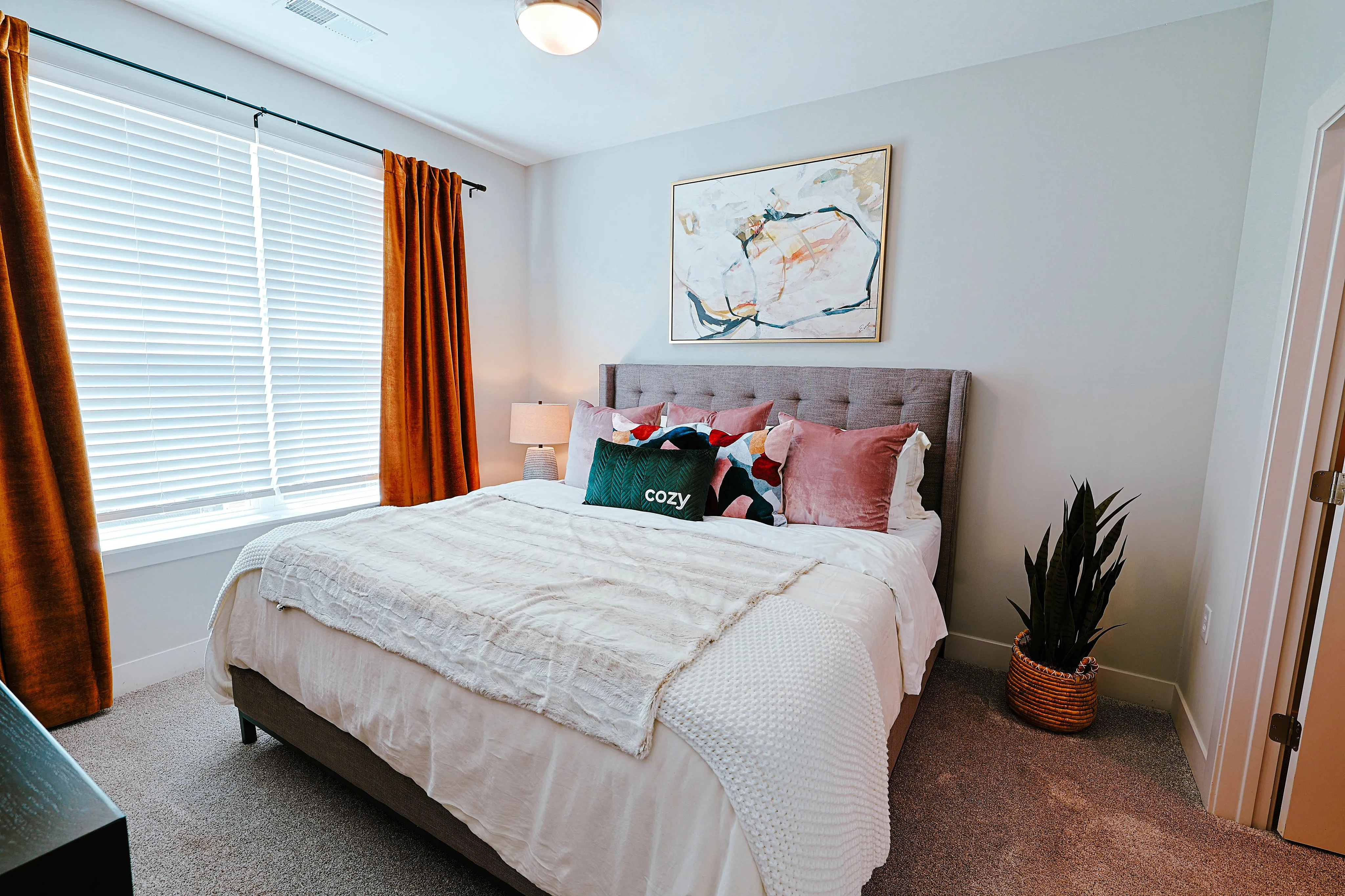 Inviting bedroom with elegant decor, featuring a plush bed adorned with vibrant pillows, large window with warm curtains, and a stylish abstract painting for a touch of sophistication.
