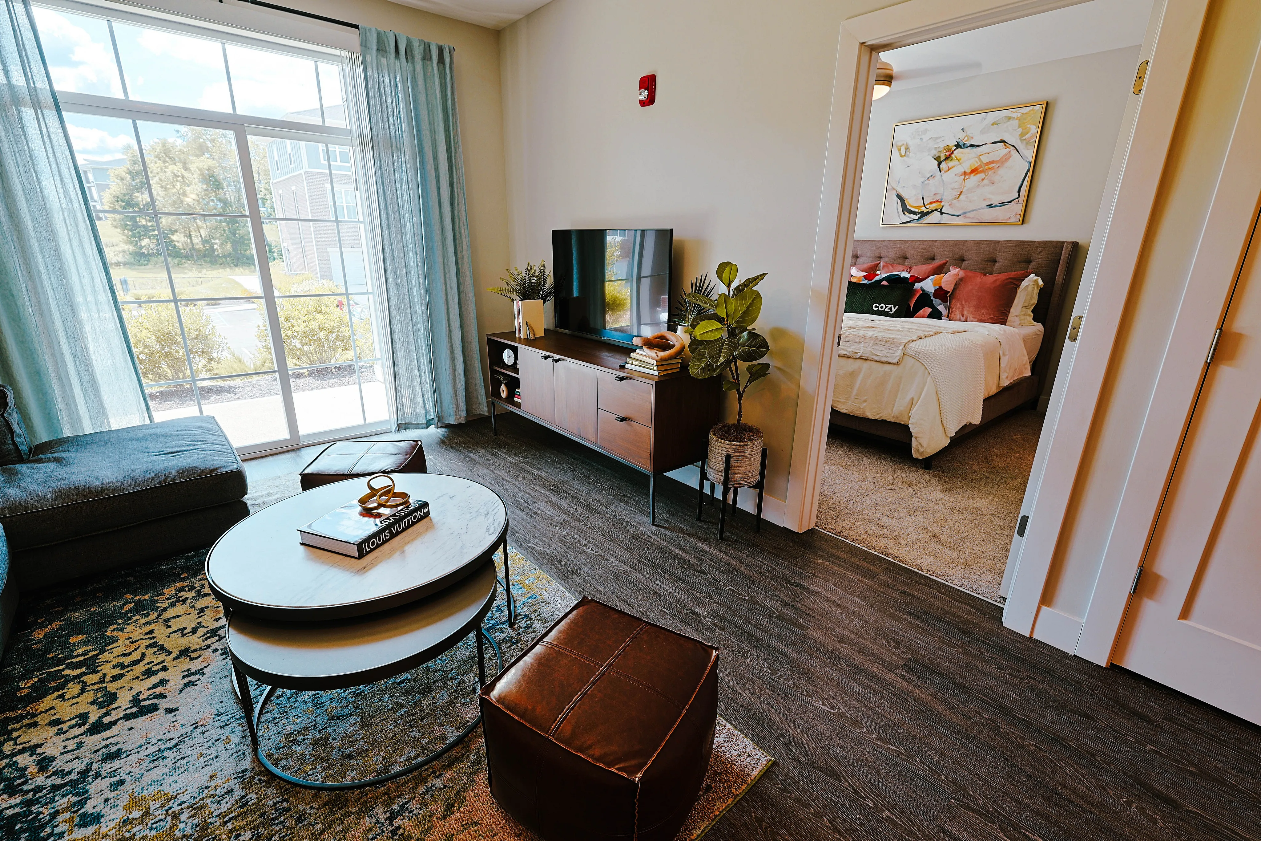 Bright living room with stylish decor, featuring a large window, comfortable seating, and a glimpse into a cozy, inviting bedroom.