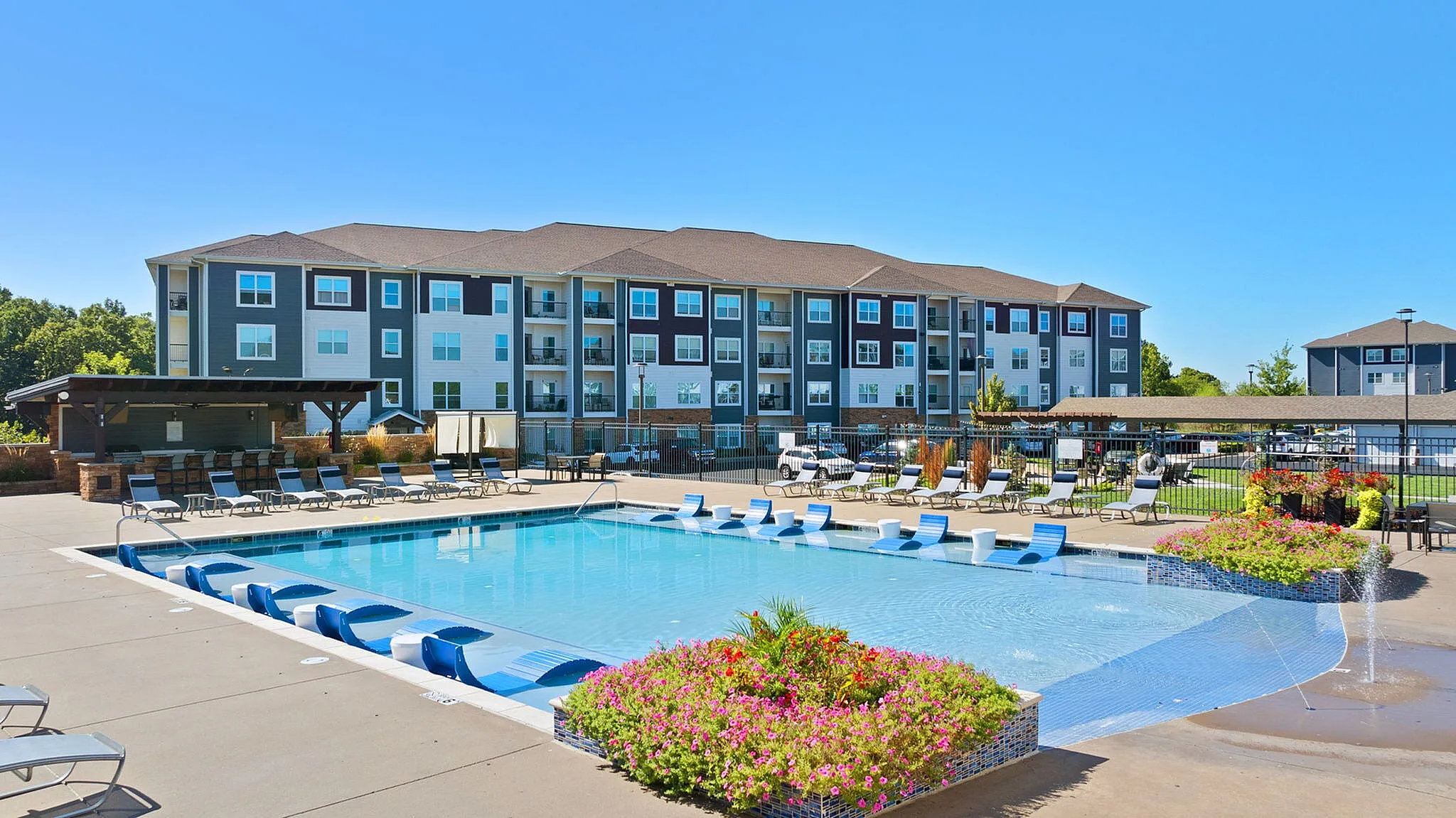 Luxurious apartment complex with a sparkling outdoor pool, modern loungers, and vibrant landscaping under a clear blue sky.
