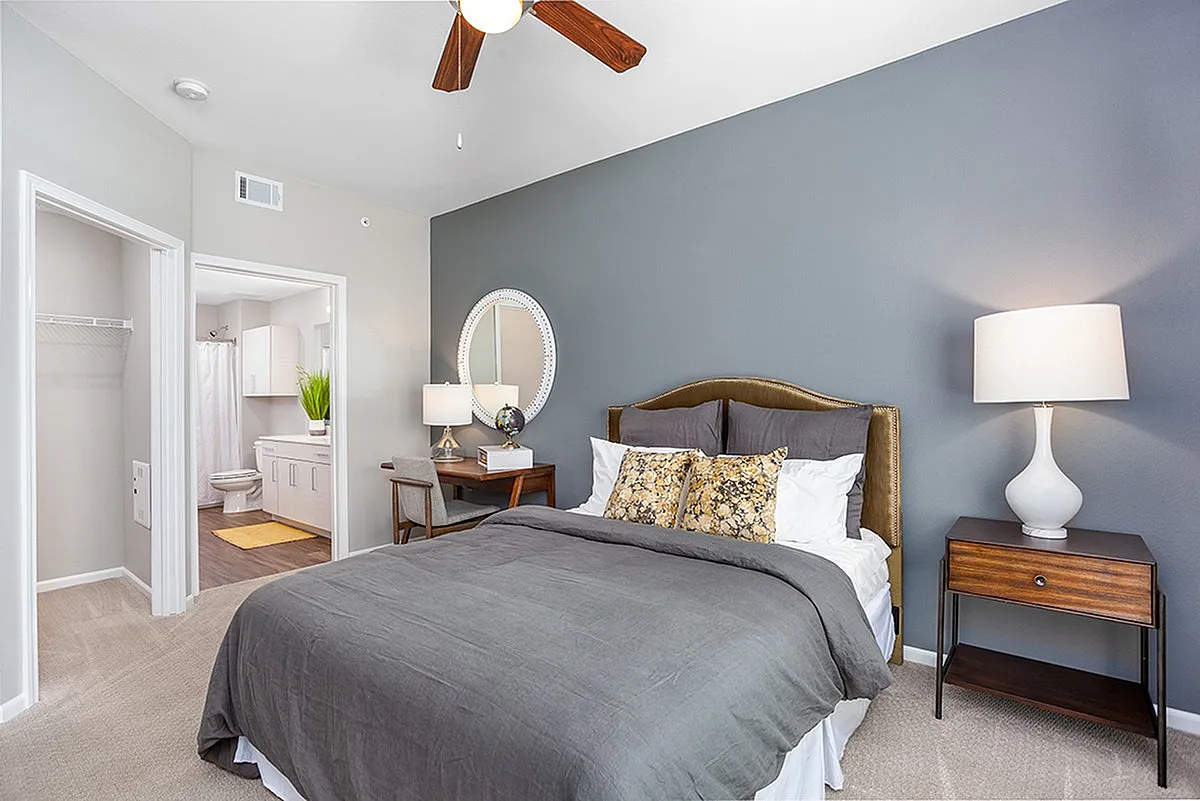 Elegant bedroom featuring a plush bed with stylish throw pillows, a wooden nightstand with a modern lamp, and a chic vanity desk. The room leads to a bright en-suite bathroom, enhancing convenience and luxury living.