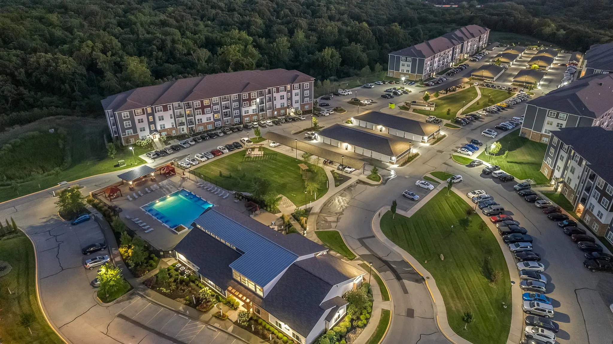 Charming aerial view of a modern apartment complex with a pristine pool, lush gardens, and ample parking, nestled in a serene woodland setting.
