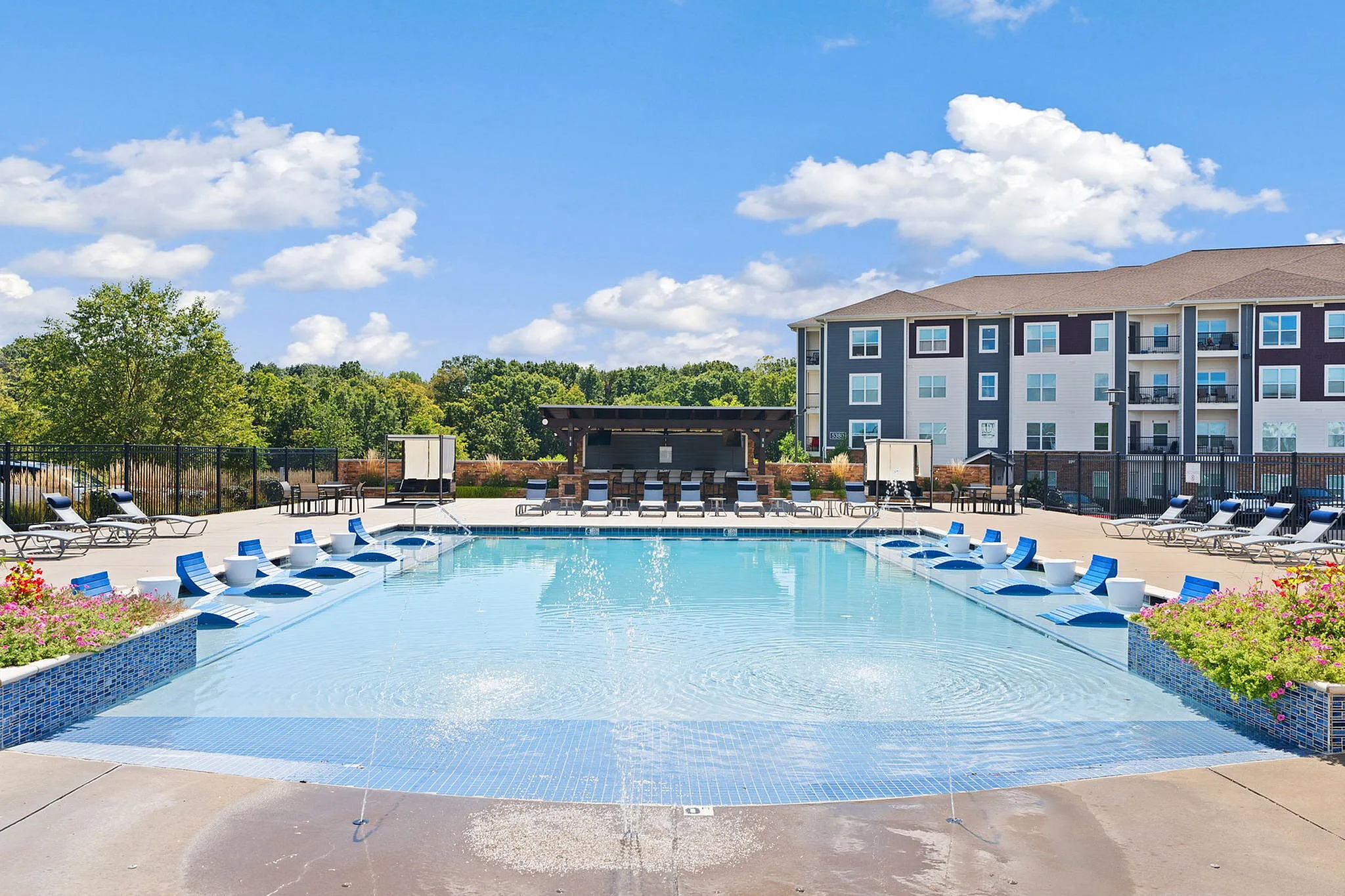 Inviting apartment complex with a large, sparkling swimming pool, surrounded by modern outdoor seating and lush landscaping under a bright blue sky.