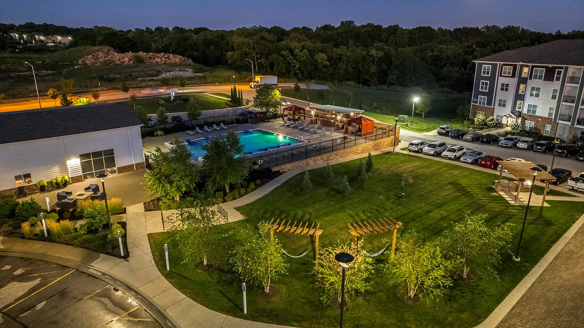 Charming apartment complex with a beautifully lit swimming pool, ample green space, and cozy pergolas, offering a serene and inviting atmosphere.