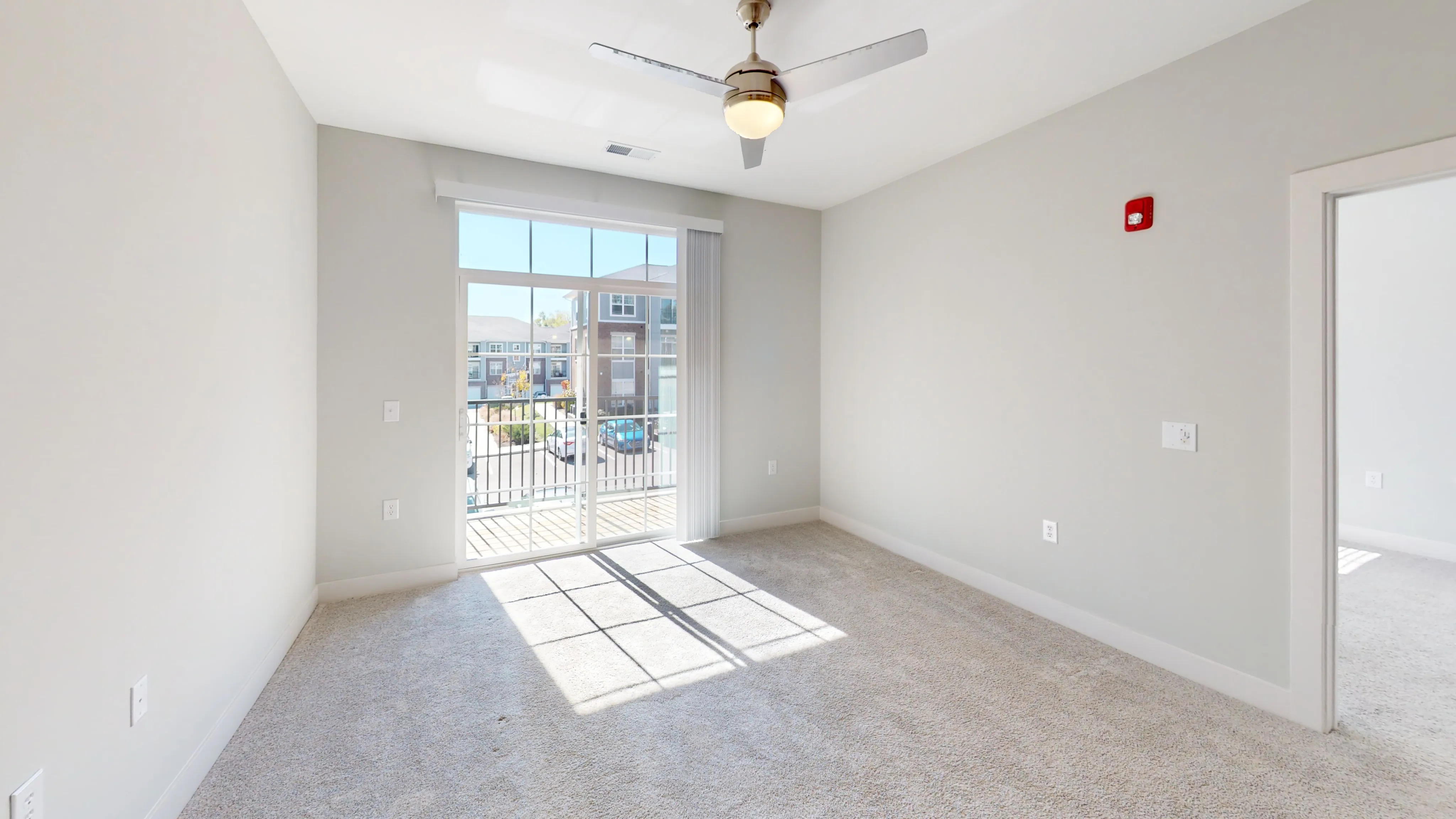 Bright and spacious bedroom with plush carpeting and a ceiling fan, featuring large windows leading to a balcony with a modern view.