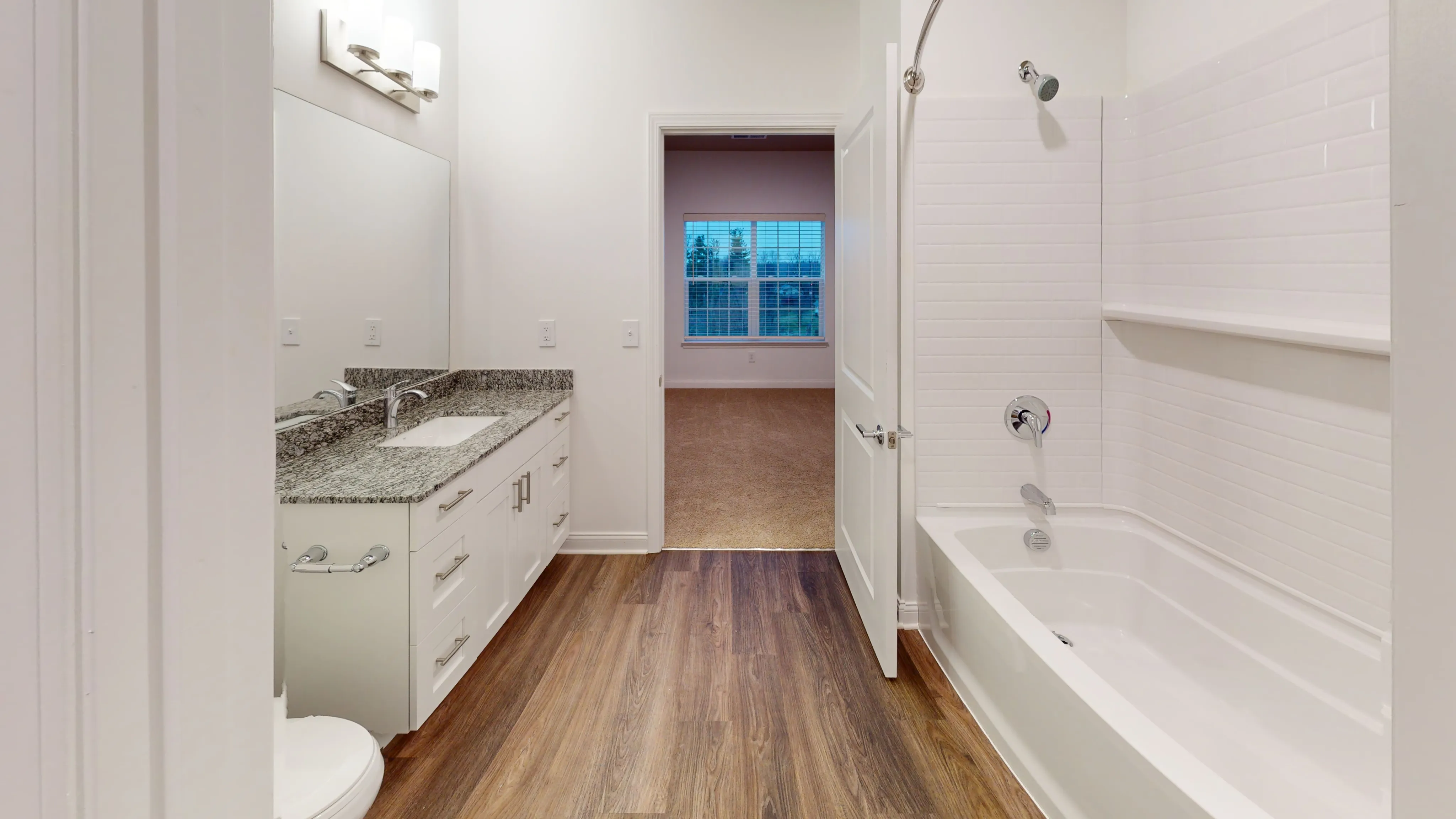 Elegant bathroom with granite countertop, modern lighting, and a spacious bathtub leading to a well-lit room.