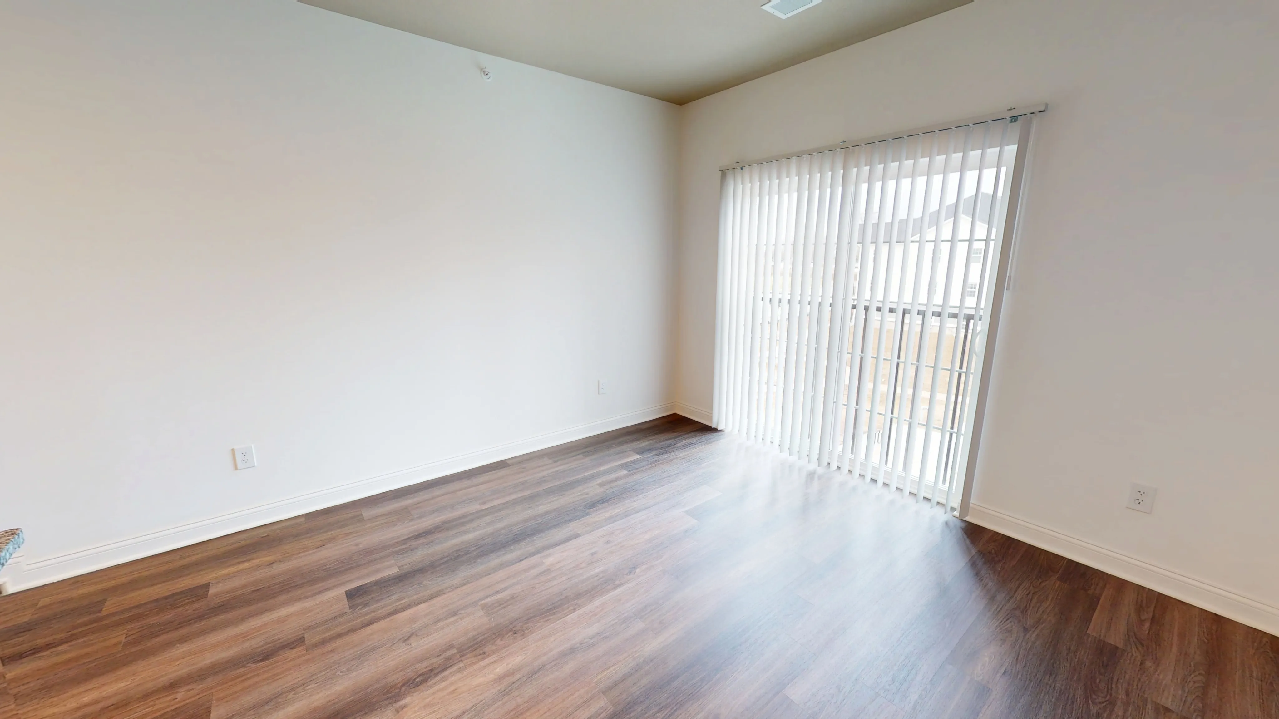 Bright room with elegant wood flooring and large window with vertical blinds, offering natural light and a welcoming atmosphere.