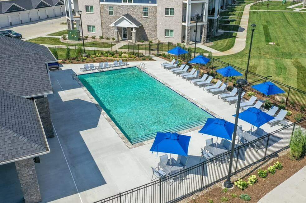 Inviting apartment complex with a sparkling swimming pool, surrounded by sleek lounge chairs and vibrant blue umbrellas, set amidst lush green landscaping.