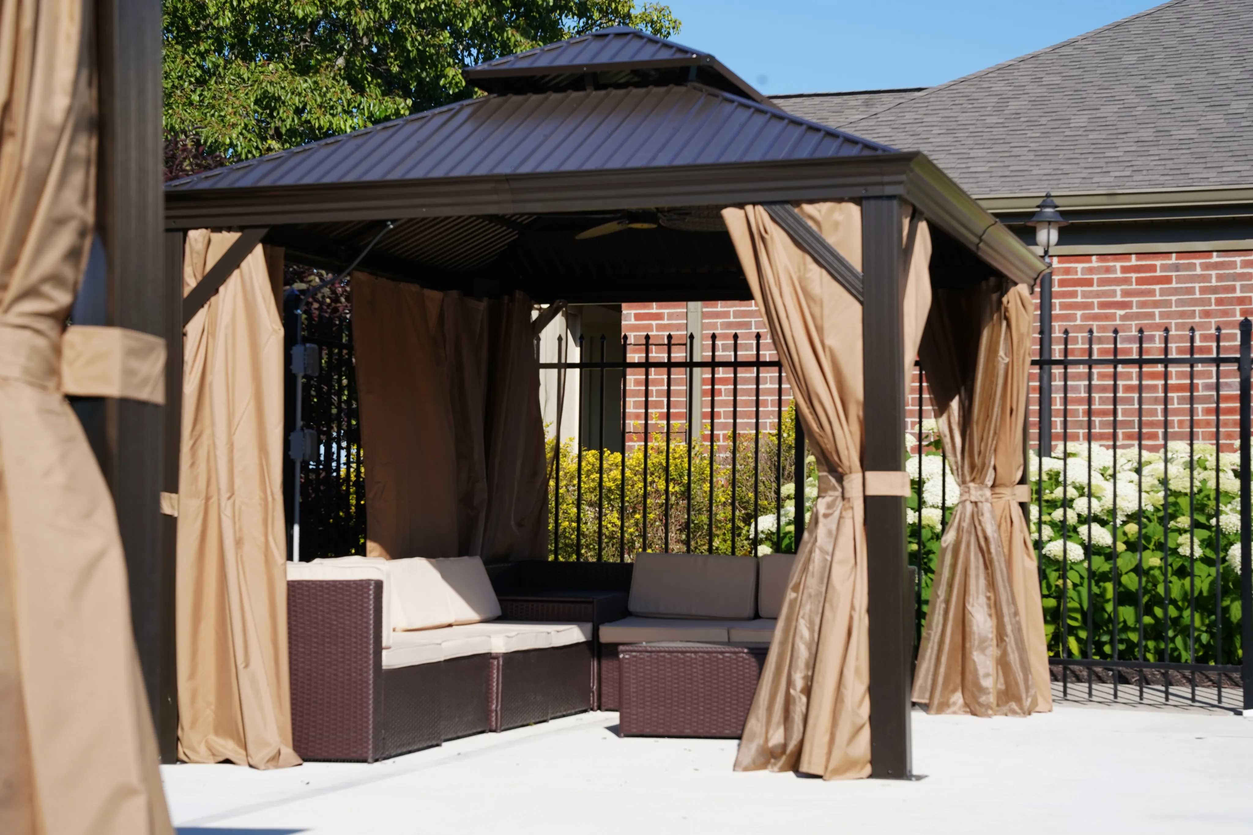 Charming outdoor gazebo with comfortable wicker seating and tan curtains, set against a lush garden backdrop for serene relaxation.