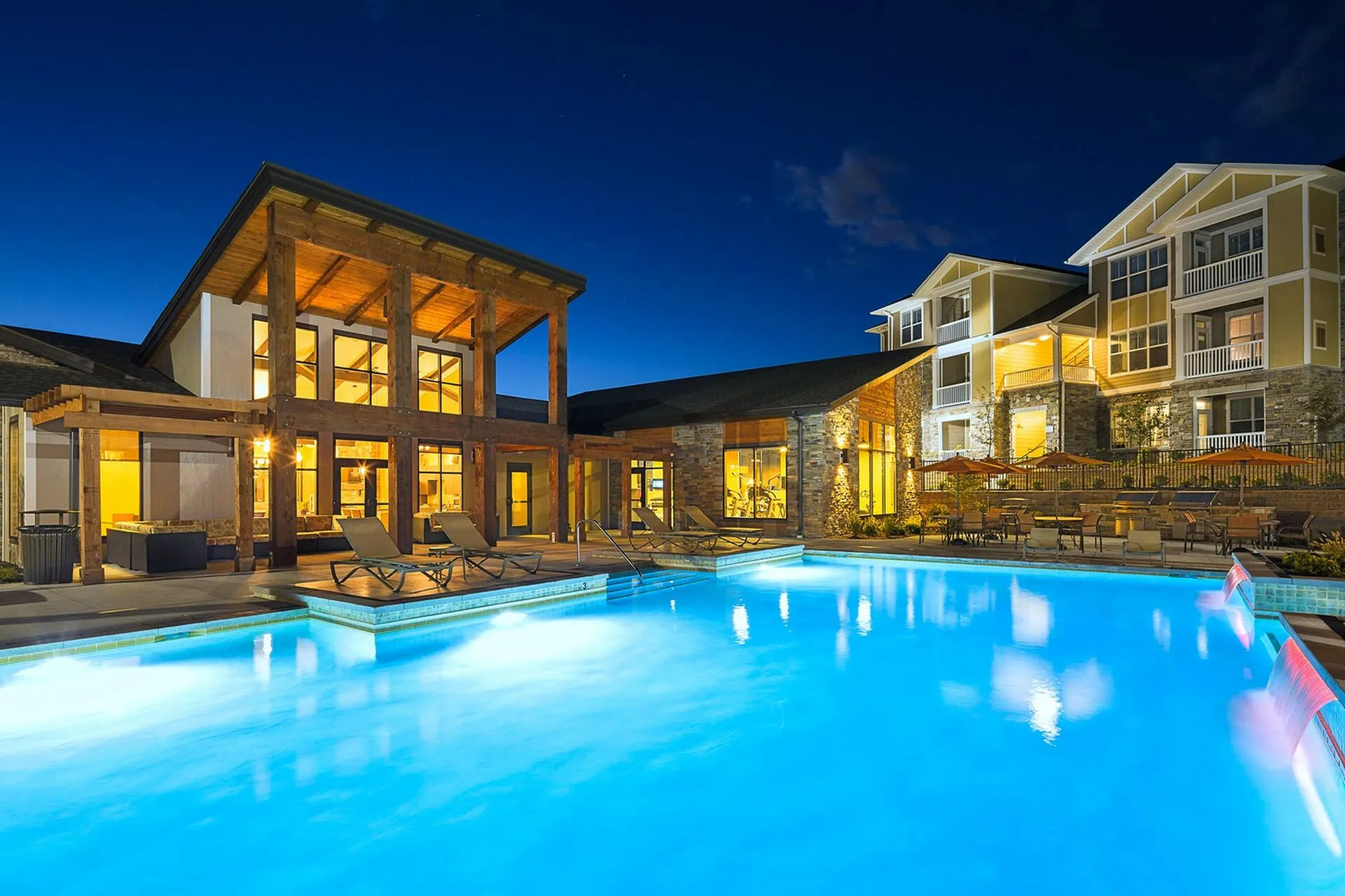 Luxurious apartment complex featuring a beautifully illuminated swimming pool at night, with elegant wooden architecture and spacious balconies.