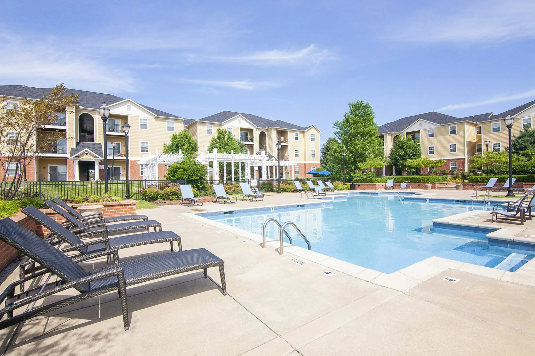Inviting apartment complex with a sparkling pool, lush landscaping, and modern sun loungers under a clear blue sky.