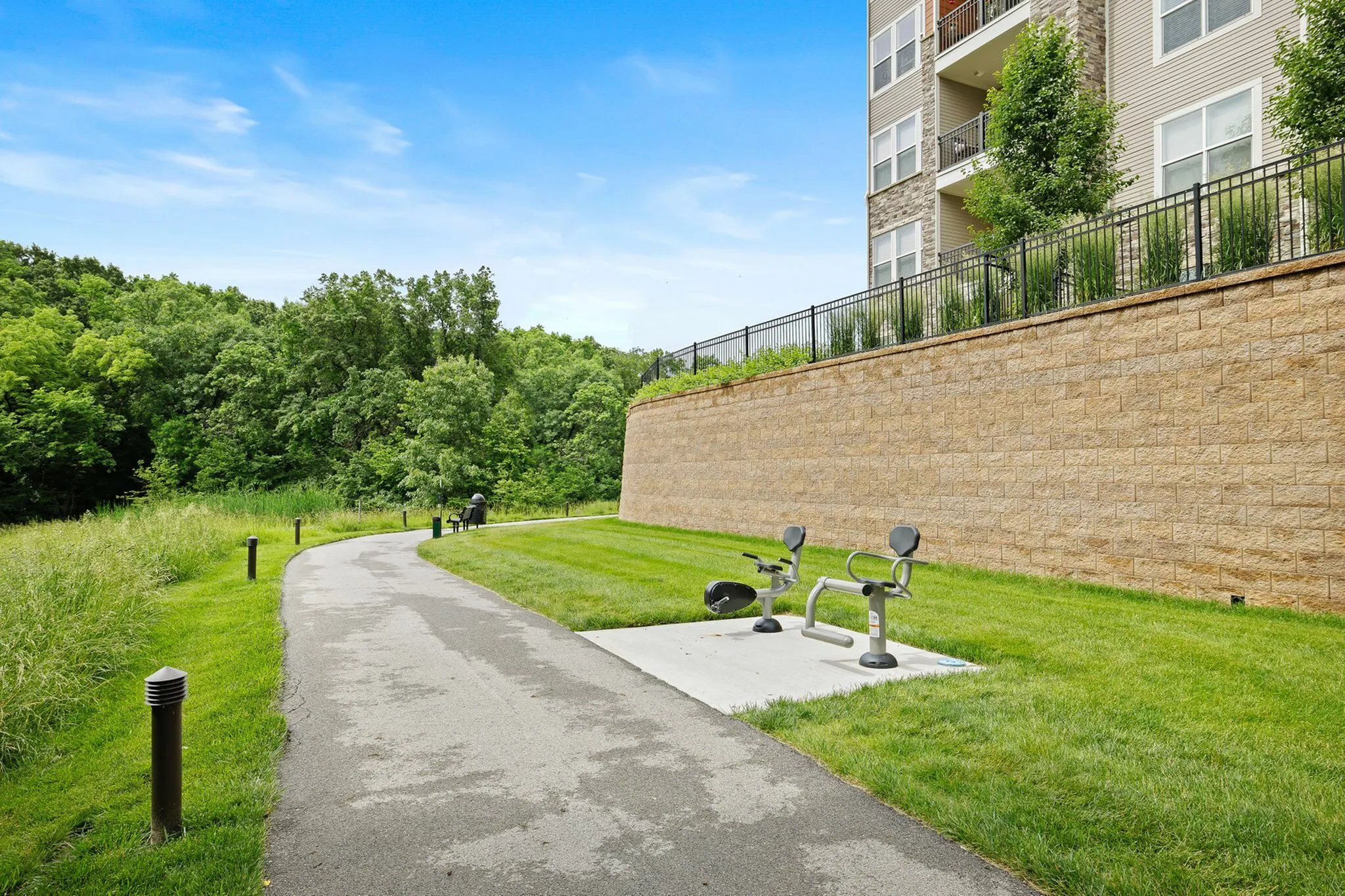 Inviting outdoor path next to modern apartment building with vibrant greenery and fitness benches under a clear blue sky.