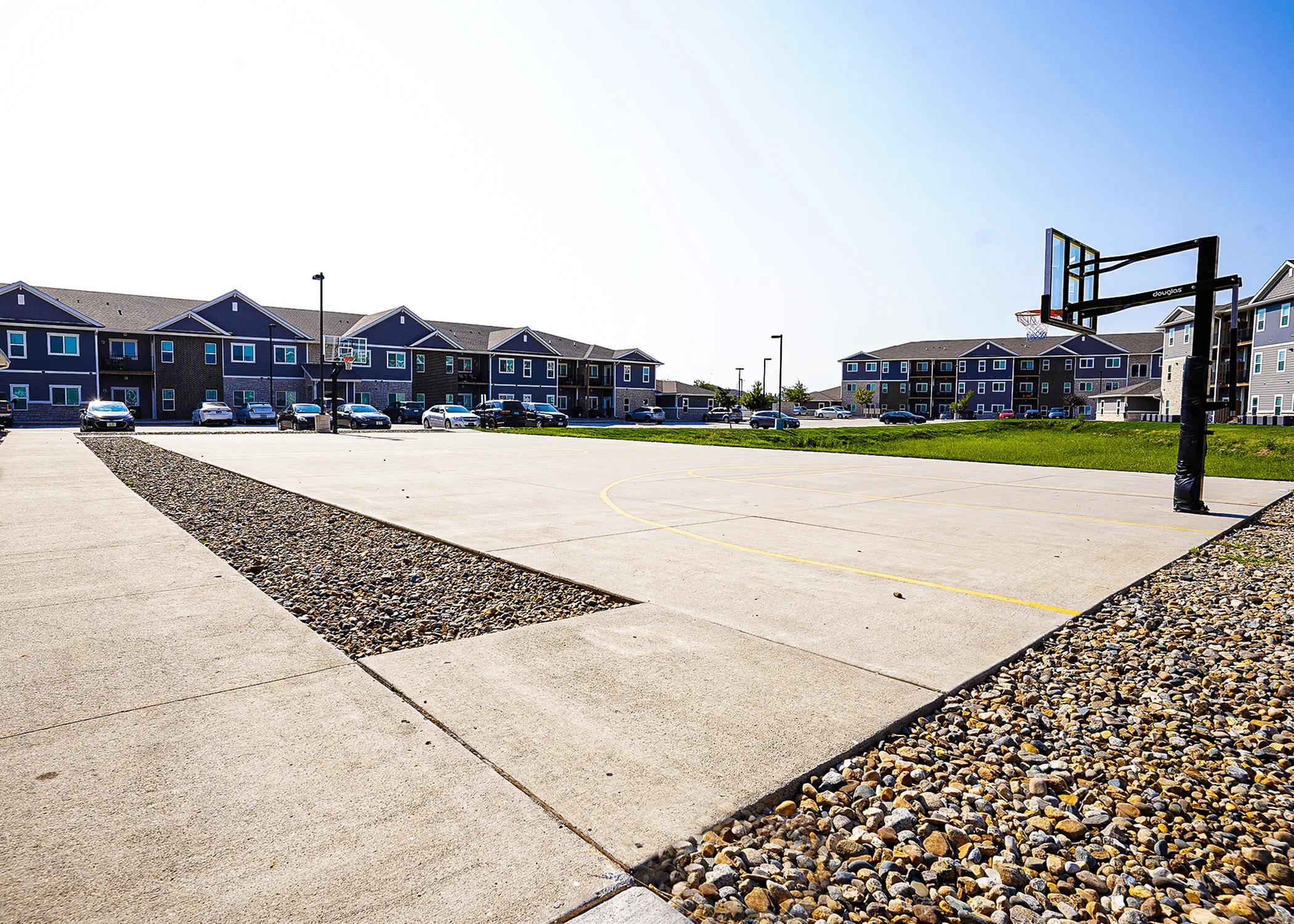 Modern apartment complex with a well-maintained outdoor basketball court, surrounded by parked cars and lush green spaces.
