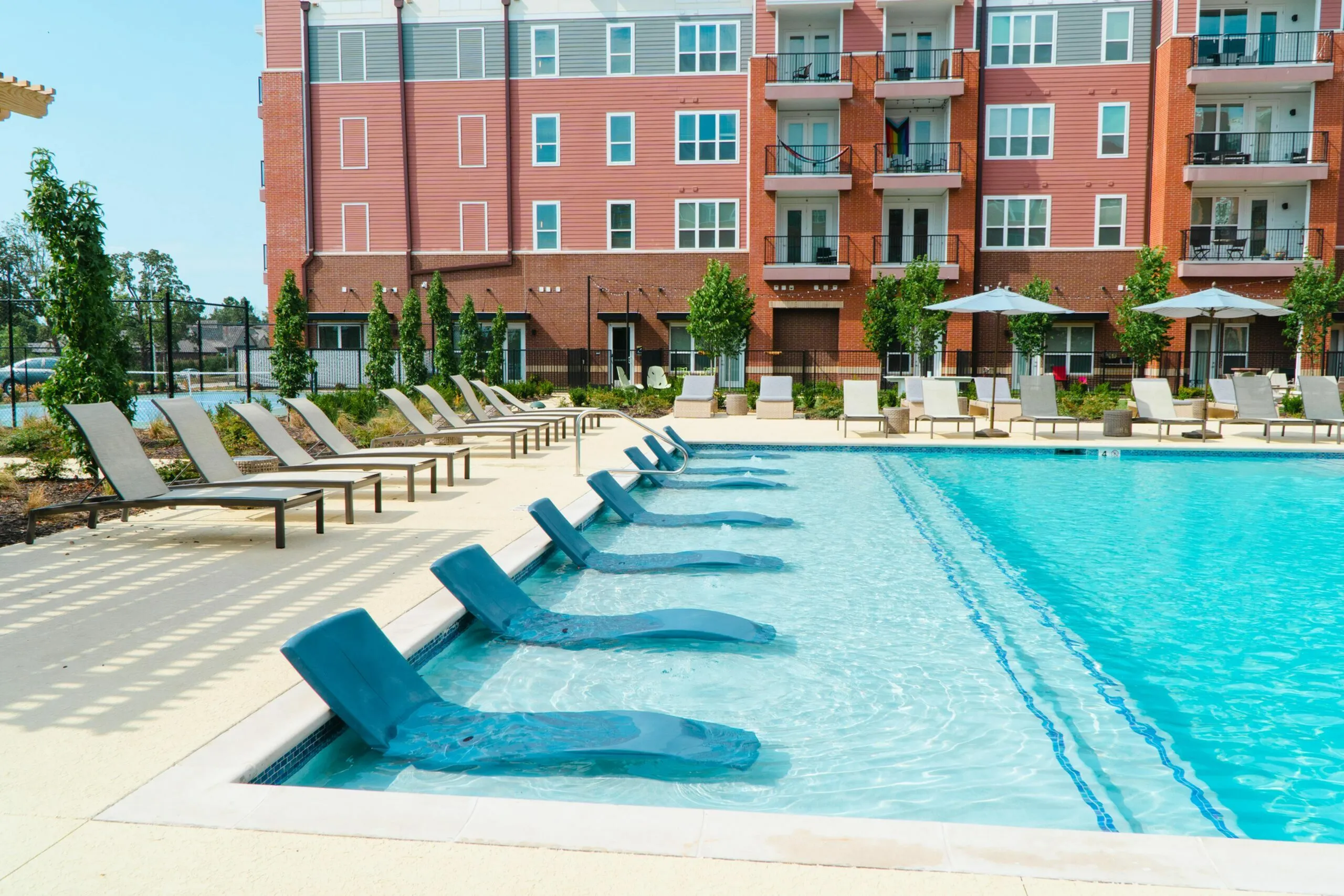 Luxurious apartment complex featuring a sparkling pool with stylish lounge chairs and parasol-covered seating, perfect for relaxation and leisure.