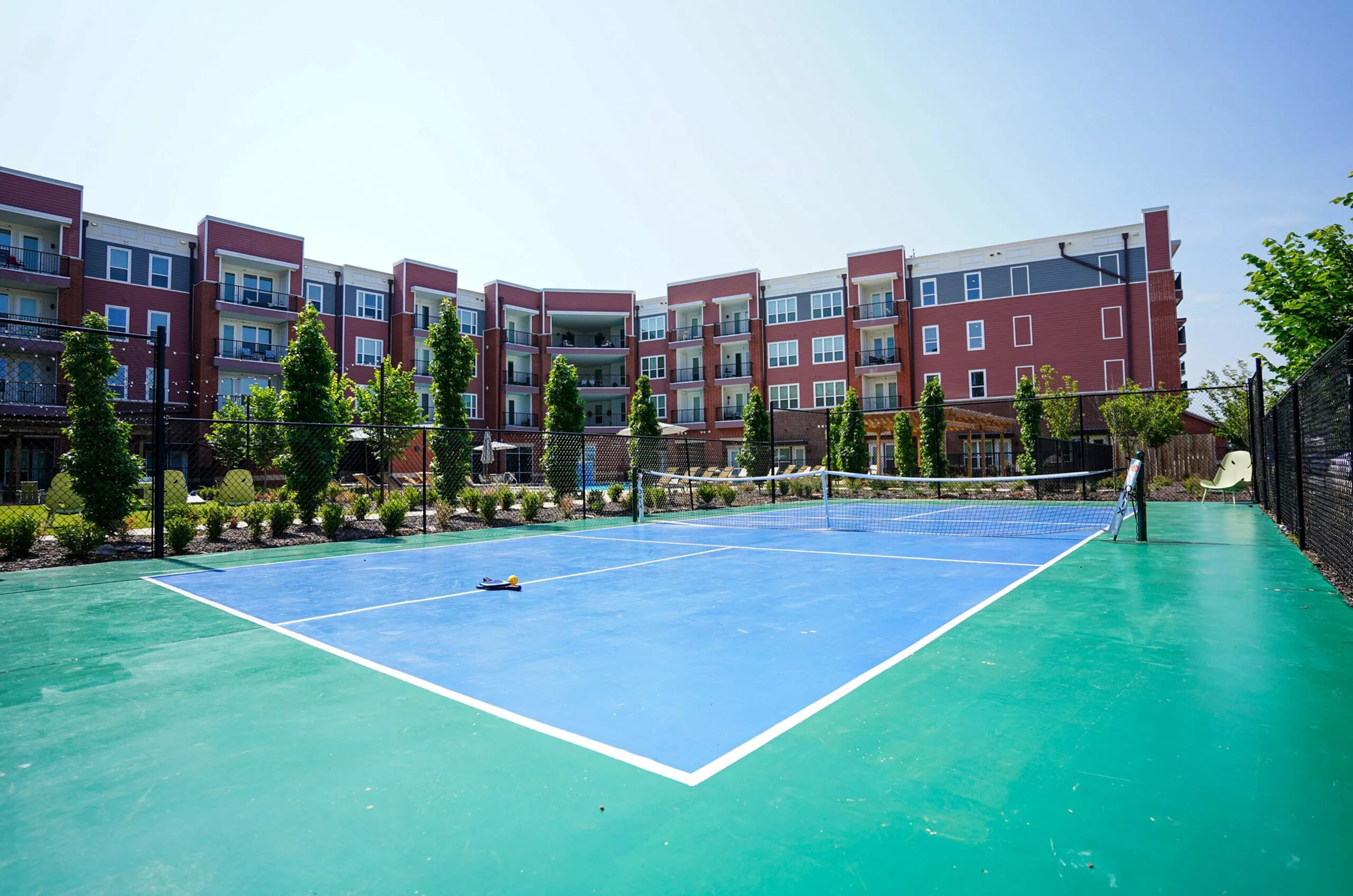Modern apartment complex with balconies overlooking a vibrant blue tennis court, surrounded by lush greenery.
