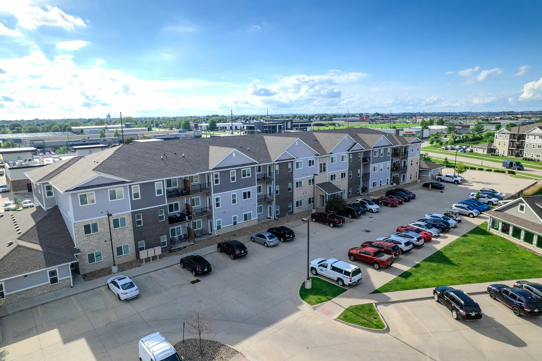 Modern apartment complex with ample parking and beautiful surrounding greenery under a bright blue sky.