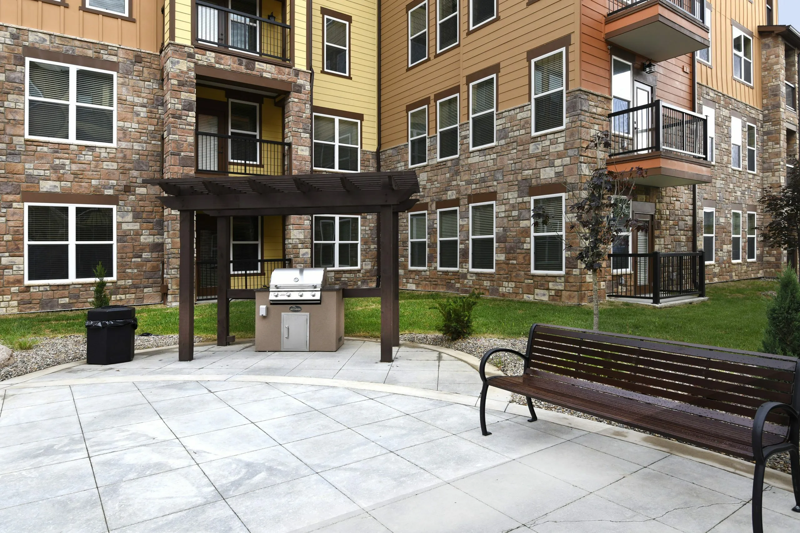 Charming apartment complex exterior featuring a stylish patio with a grill, wooden pergola, and a comfortable bench surrounded by lush greenery.