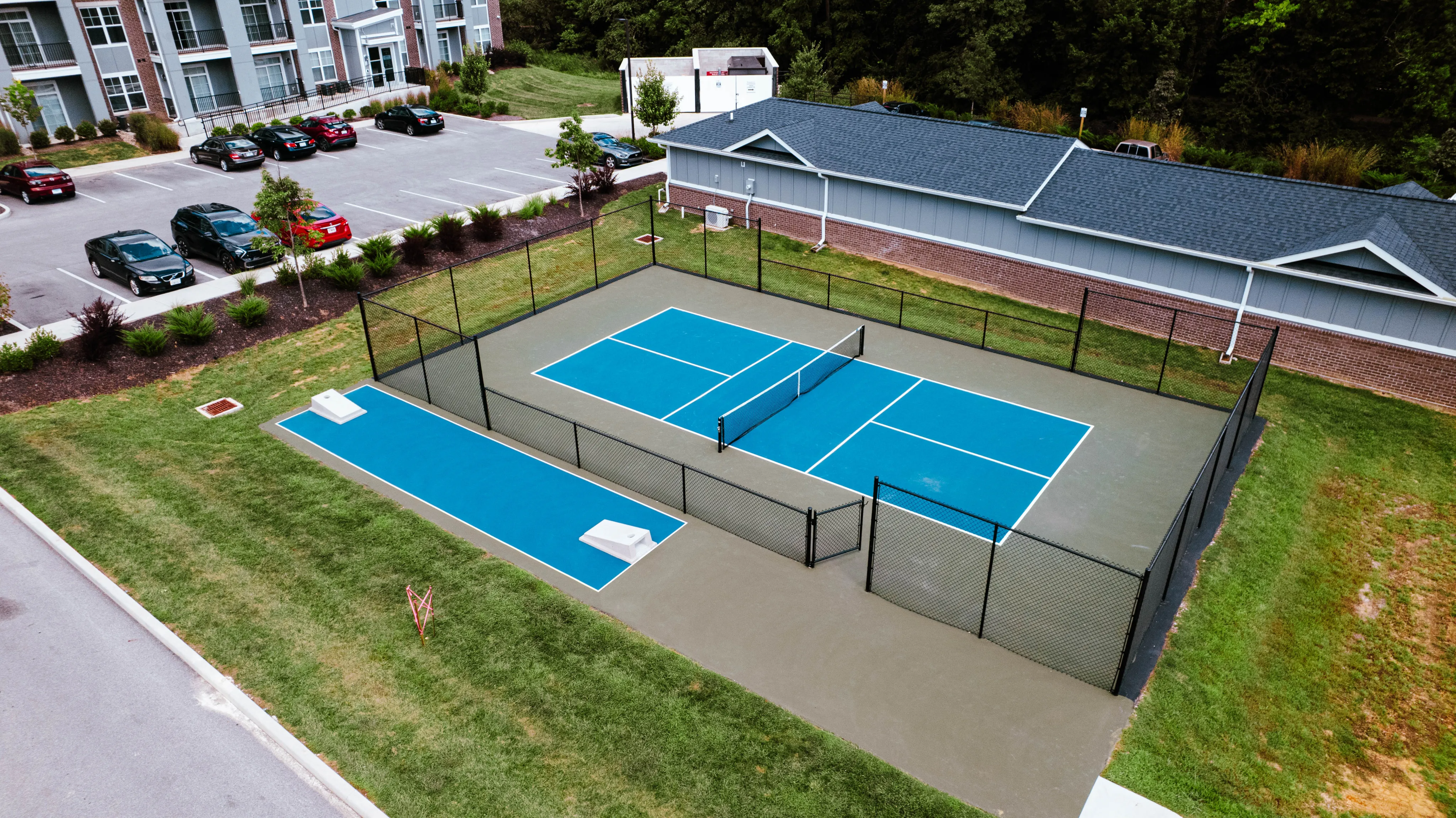 Vibrant aerial view of modern apartment complex with a tennis court, surrounded by lush greenery and ample parking.