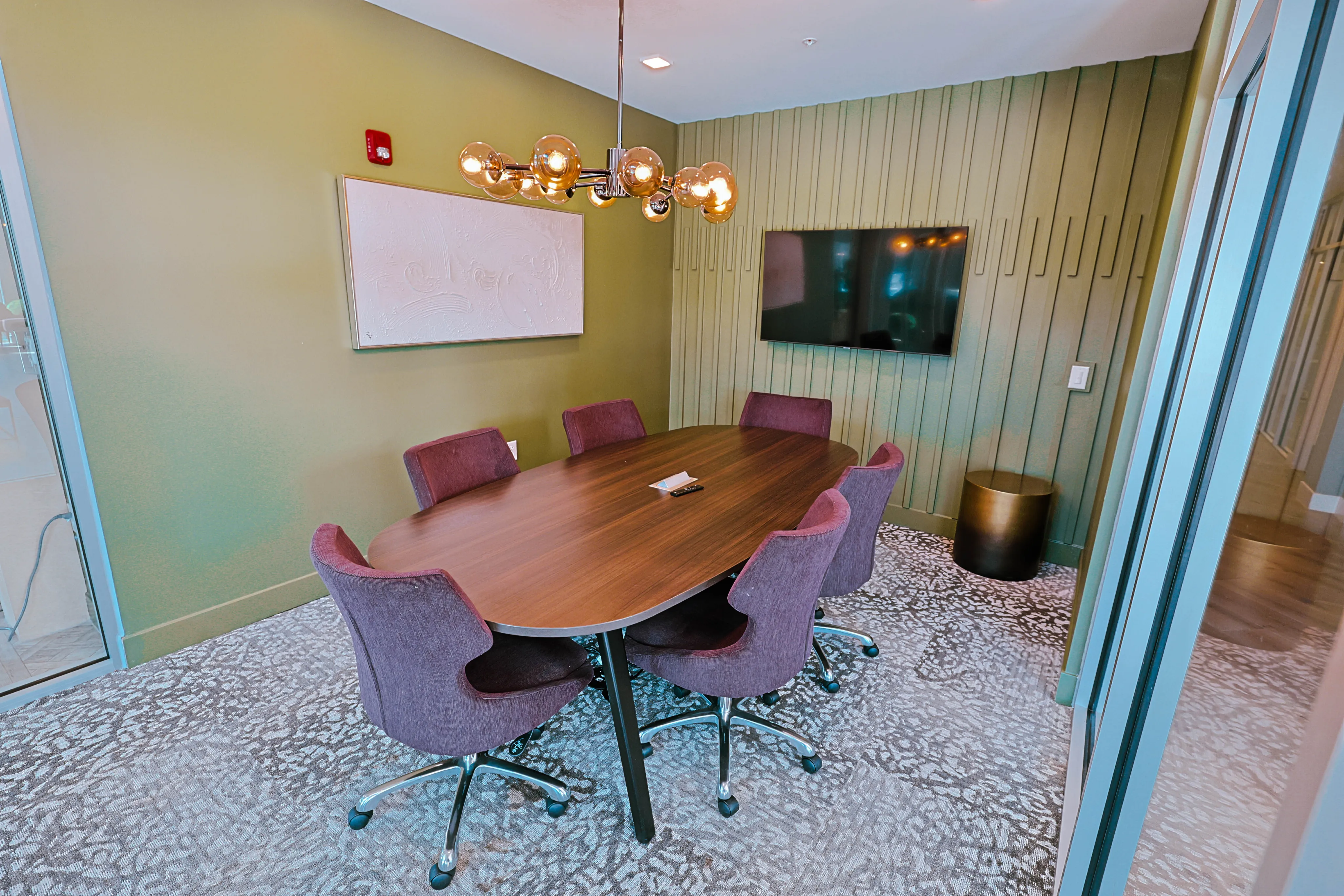 Chic meeting room featuring a large oval wooden table, plush purple chairs, modern light fixture, and sleek wall-mounted TV.