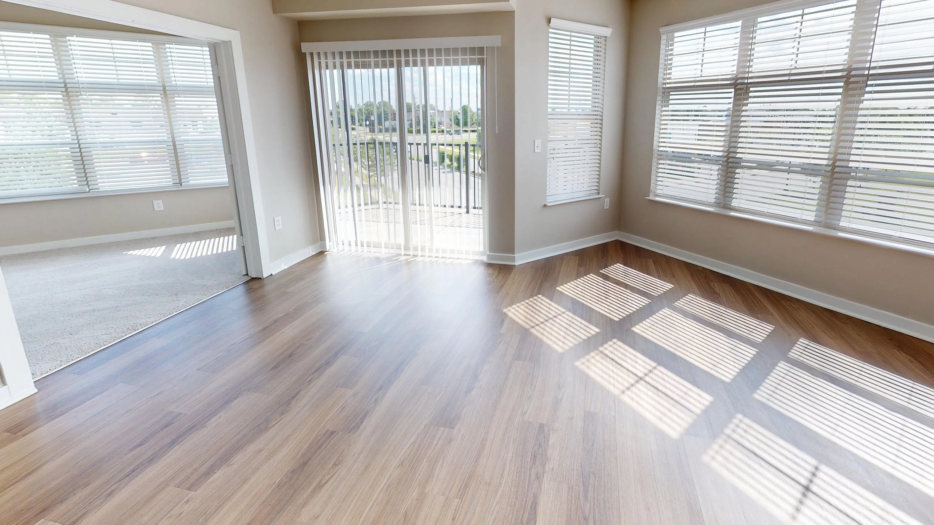 Bright and airy living space featuring large windows with chic blinds, elegant wood flooring, and modern ceiling fan, offering a seamless transition to a cozy carpeted room.