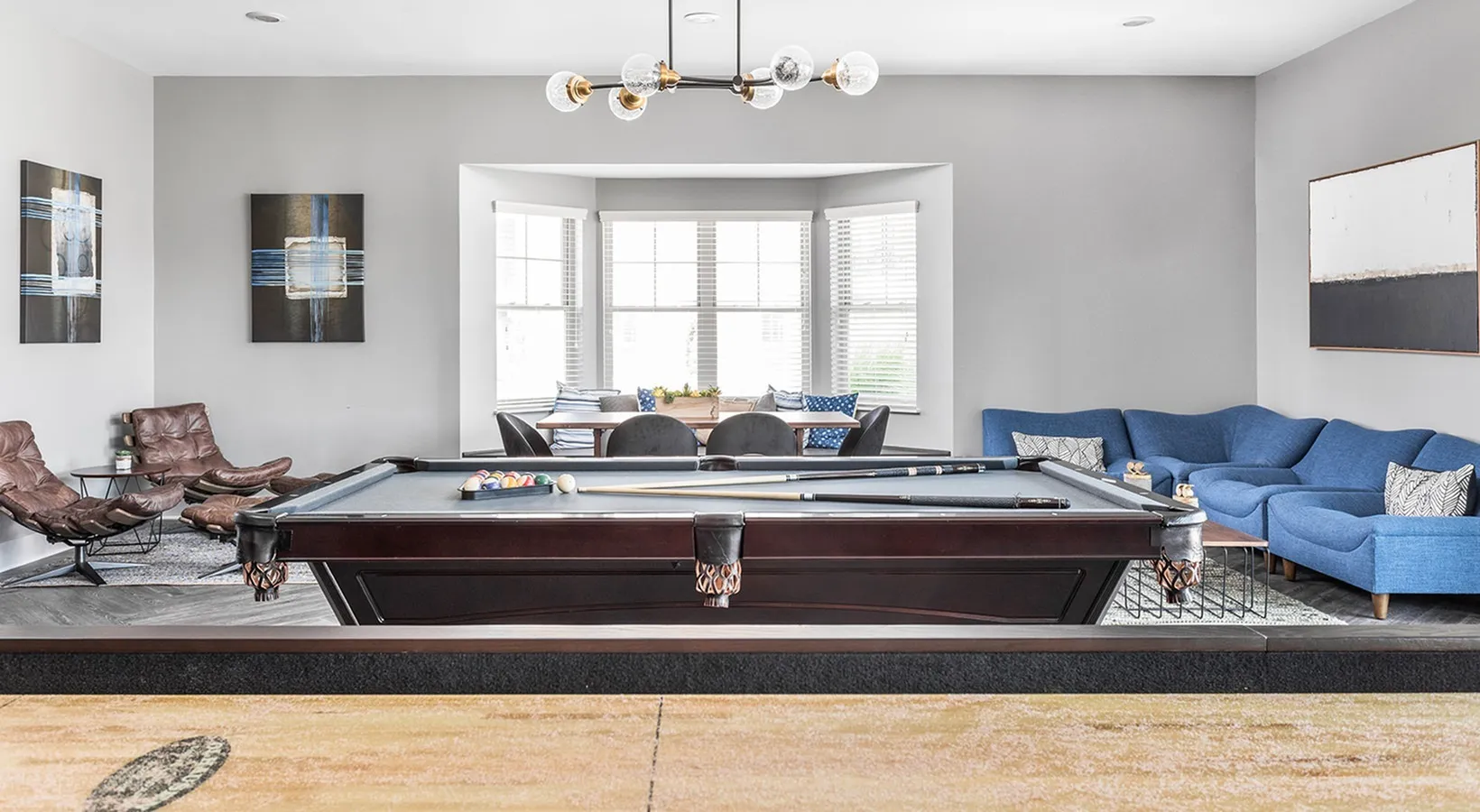 Chic lounge showcasing a pool table, modern artwork, and a cozy seating area with stylish blue couches and a sunlit dining nook.