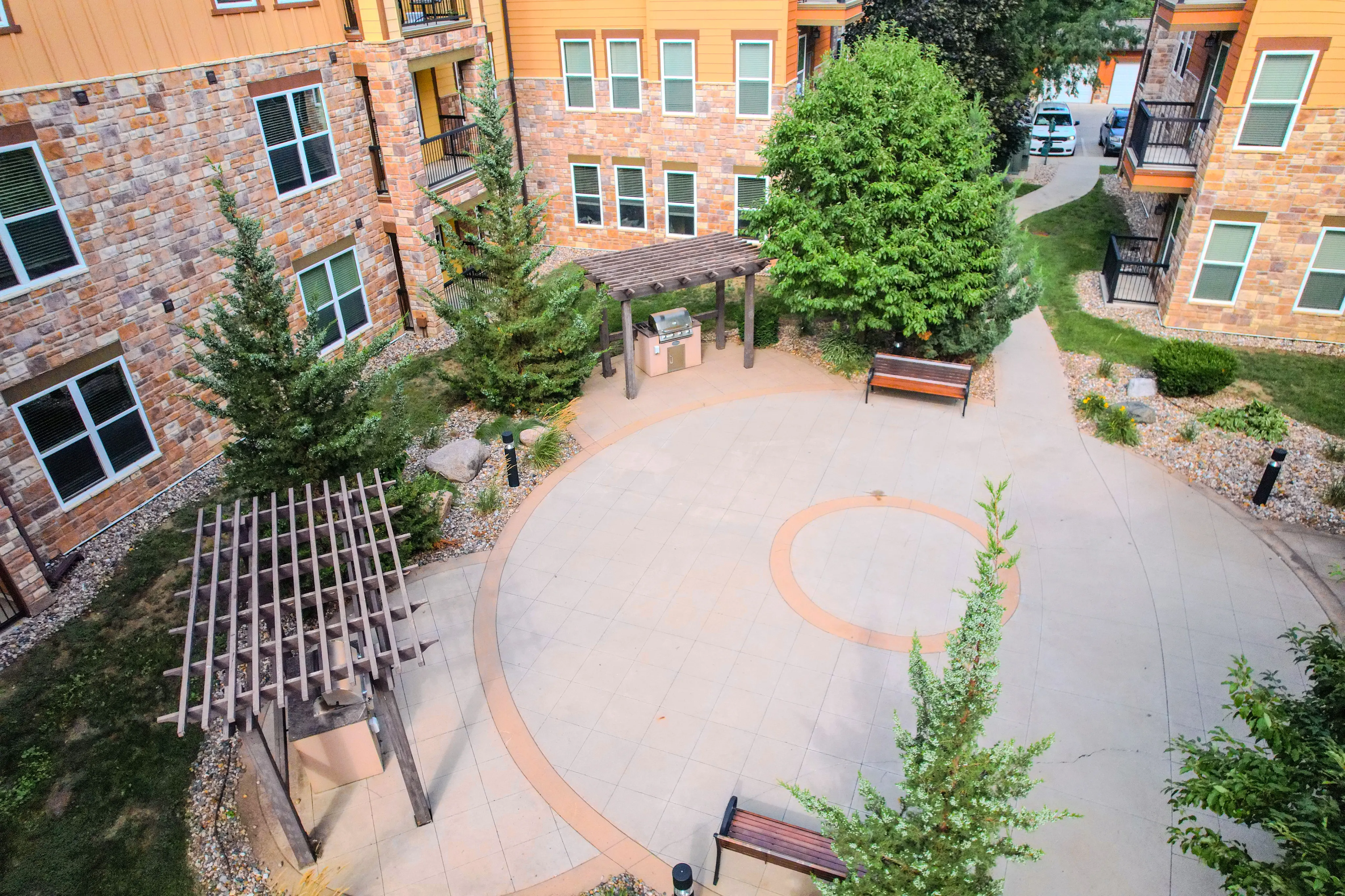 Charming landscaped courtyard featuring a circular plaza with benches, lush trees, and a cozy pergola in a brick apartment setting.