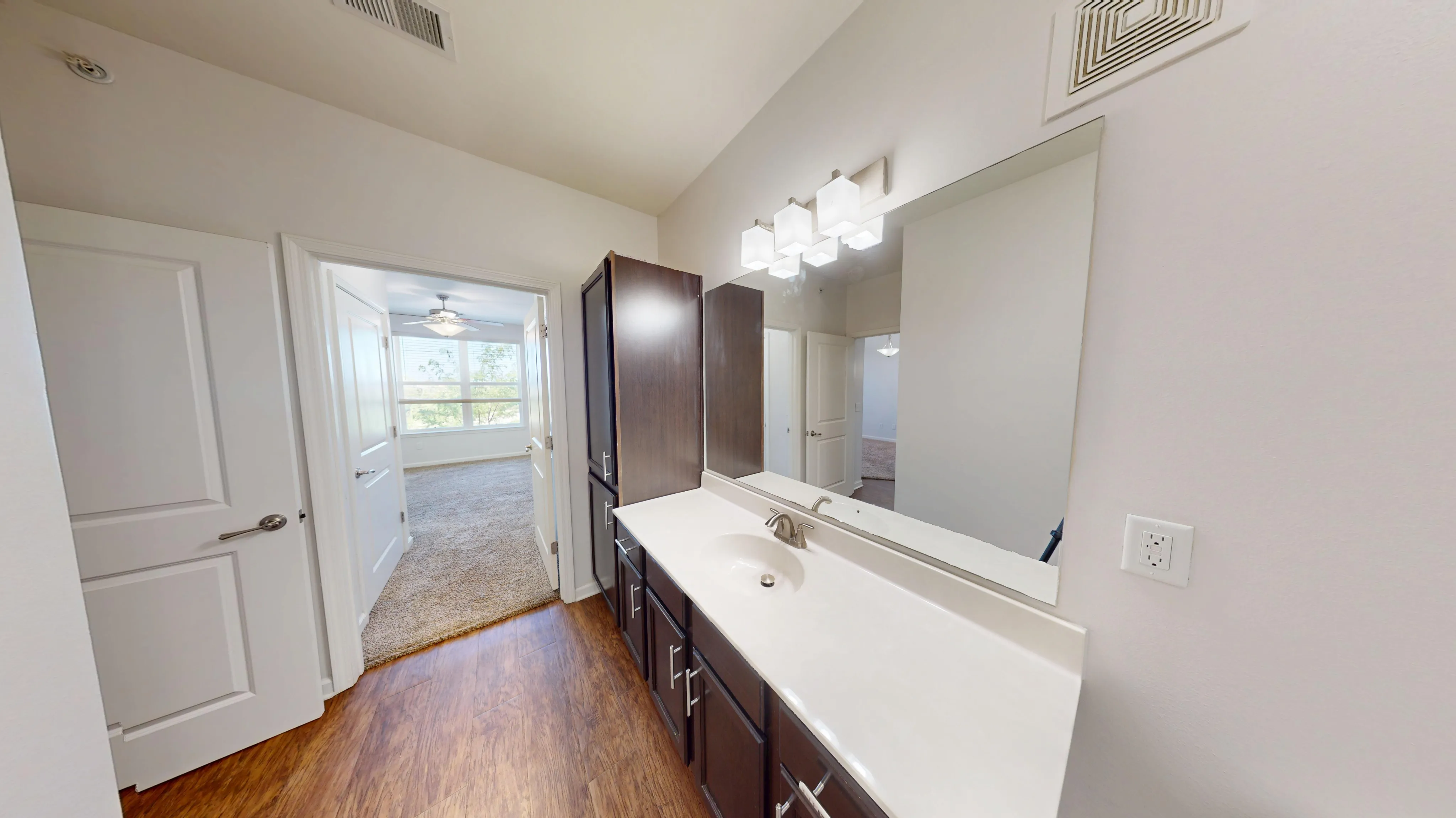 Modern bathroom with sleek dark wood cabinetry, elegant basin, and a large mirror, leading to a sunlit bedroom with plush carpet flooring.