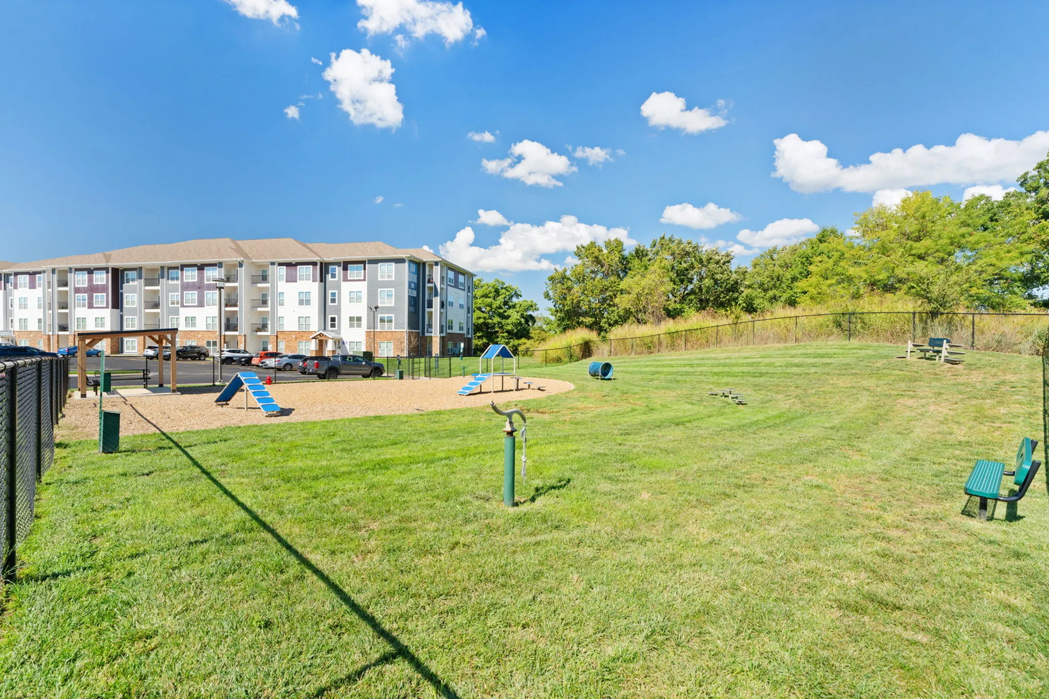 Modern apartment complex with a spacious dog park, vibrant green grass, agility equipment, and seating areas, set against a clear blue sky backdrop.